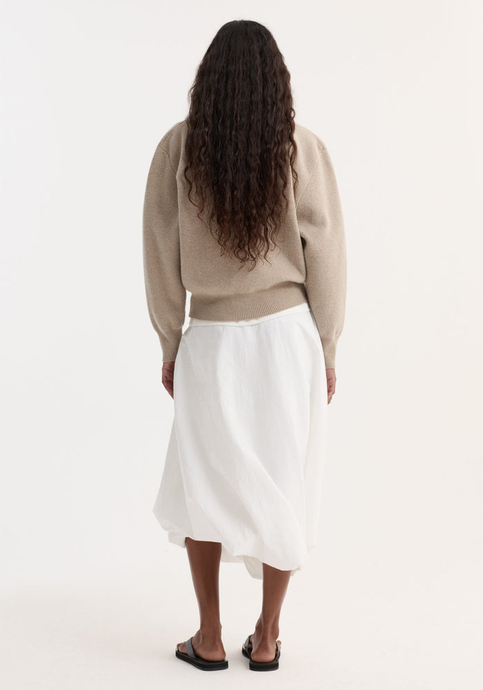 Person in beige Wool Cashmere Polo with a structured collar and three-button closure, paired with a white skirt and black slide sandals, viewed from behind.