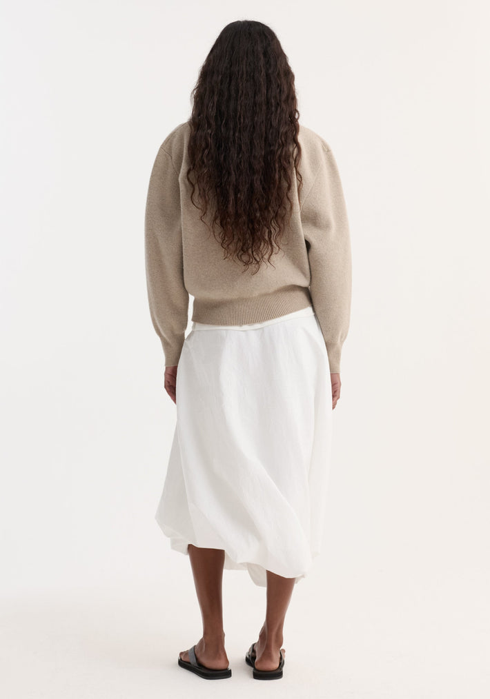 Person in beige Wool Cashmere Polo with a structured collar and three-button closure, paired with a white skirt and black slide sandals, viewed from behind.