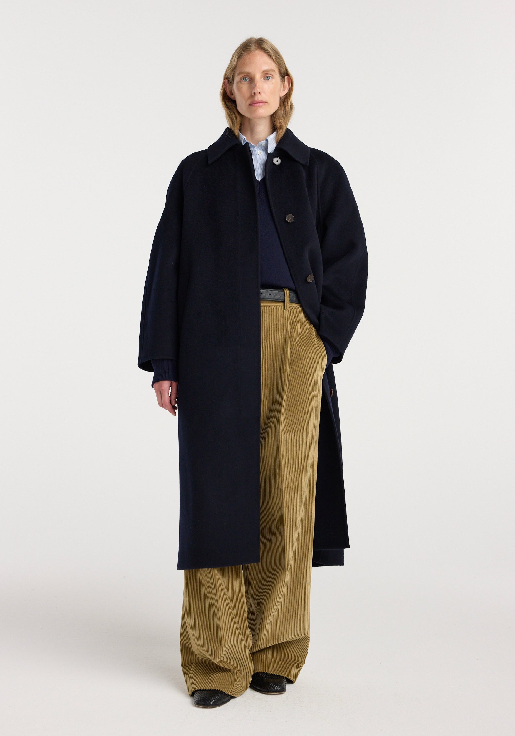 A model wearing a long double-faced wool padded coat | navy over a navy sweater and light blue shirt, paired with loose-fitting beige corduroy pants and black shoes.