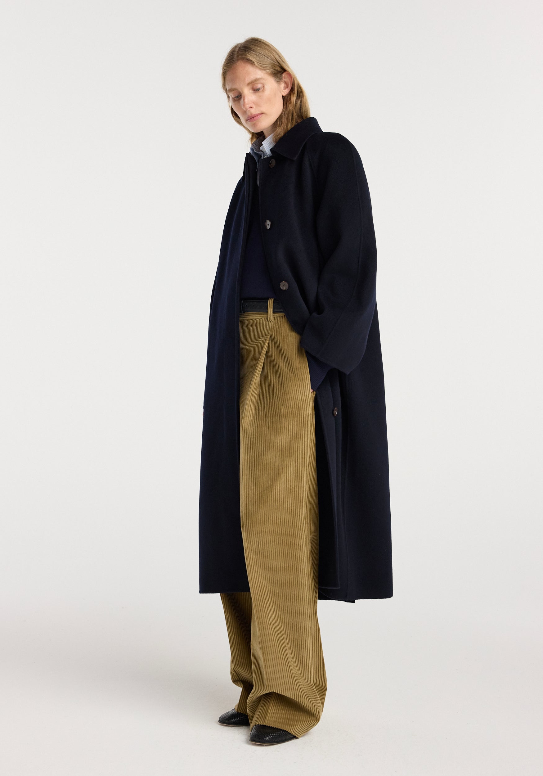 Woman wearing a navy double-faced wool padded coat with large lapels and buttons, styled with beige wide-leg corduroy pants and black shoes, standing against a plain background.
