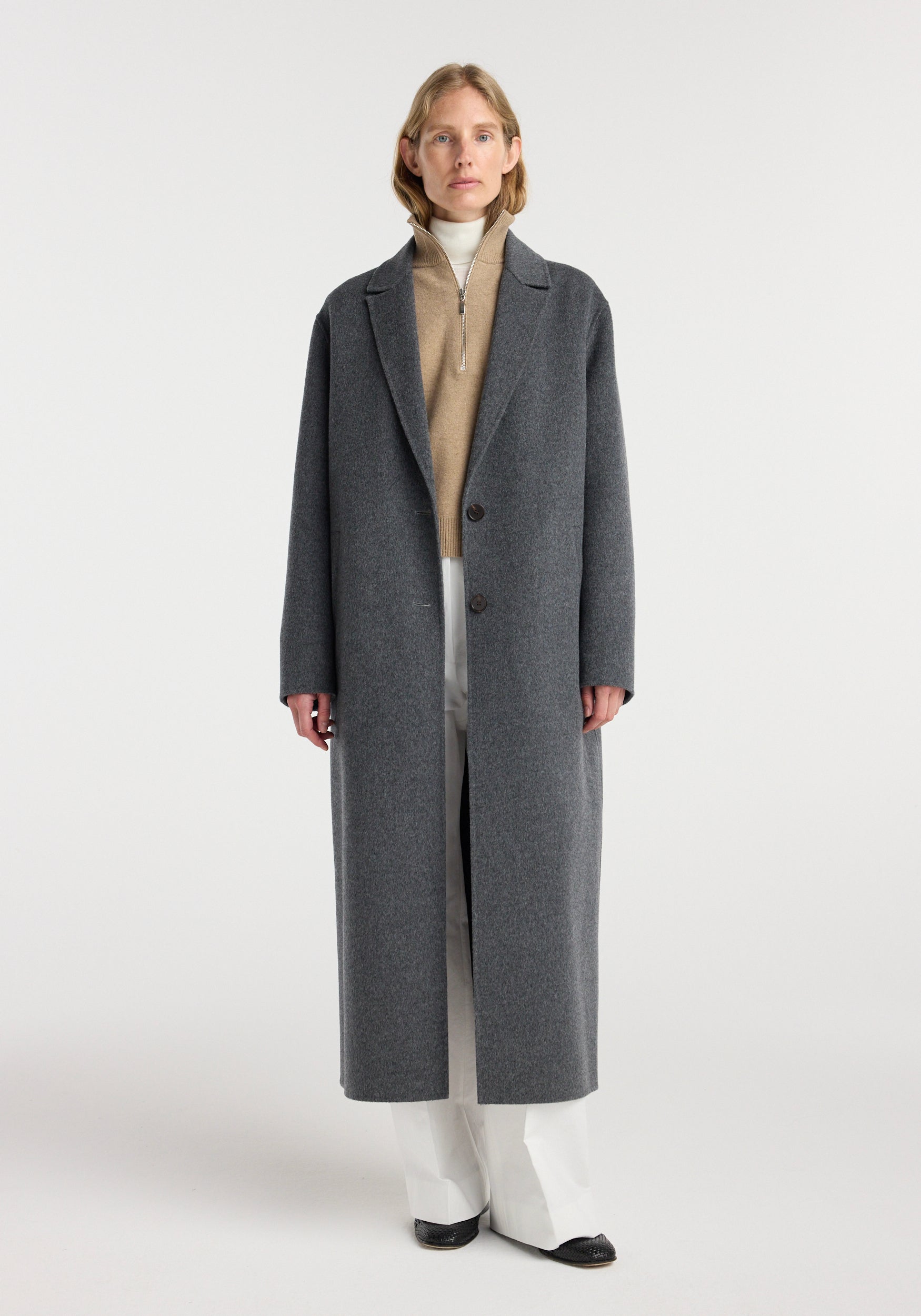 Woman wearing a long, double-faced wool long coat | dark grey melange with a beige zip sweater, white pants, and black shoes against a plain background.