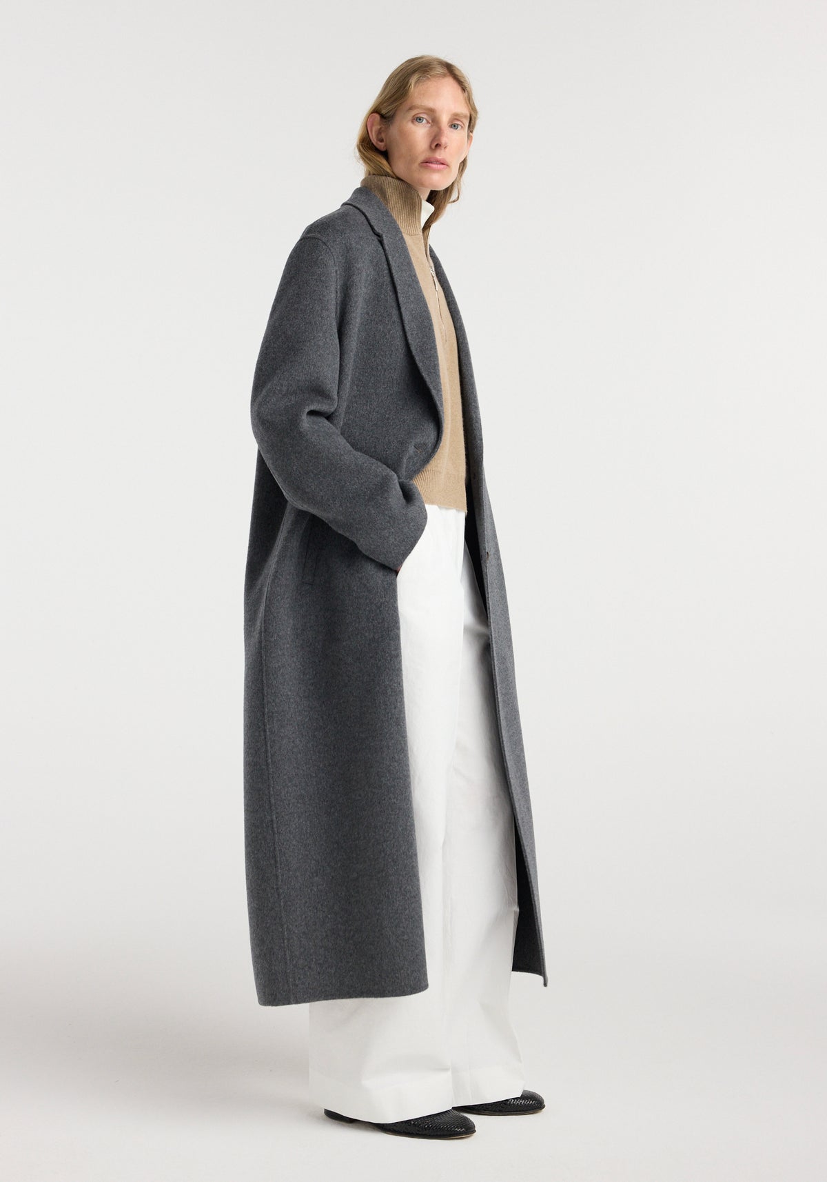 Woman wearing a dark grey melange double-faced wool long coat over a beige turtleneck sweater and wide white pants, standing against a plain light background.