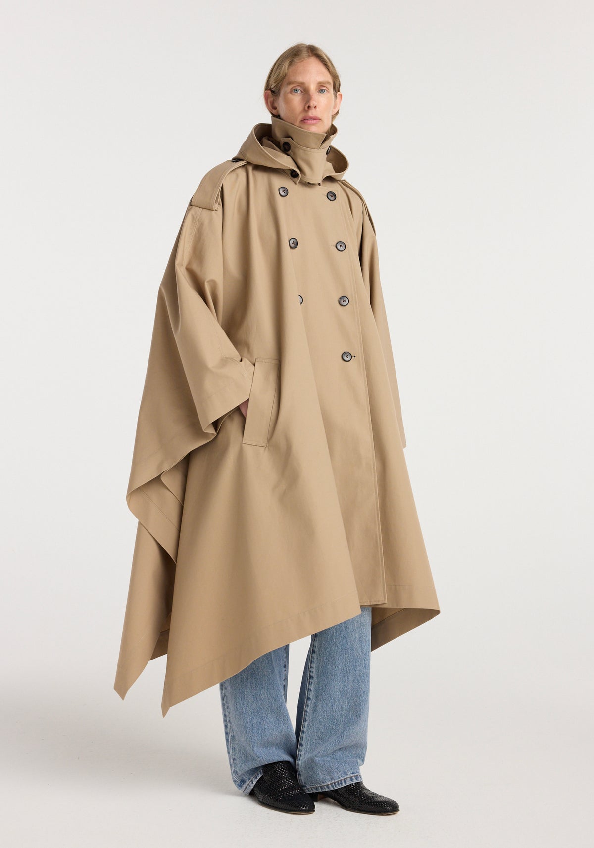 Model wearing a beige hooded cape trenchcoat with double-breasted buttons, large collar, and side pockets, paired with light blue jeans and black shoes.