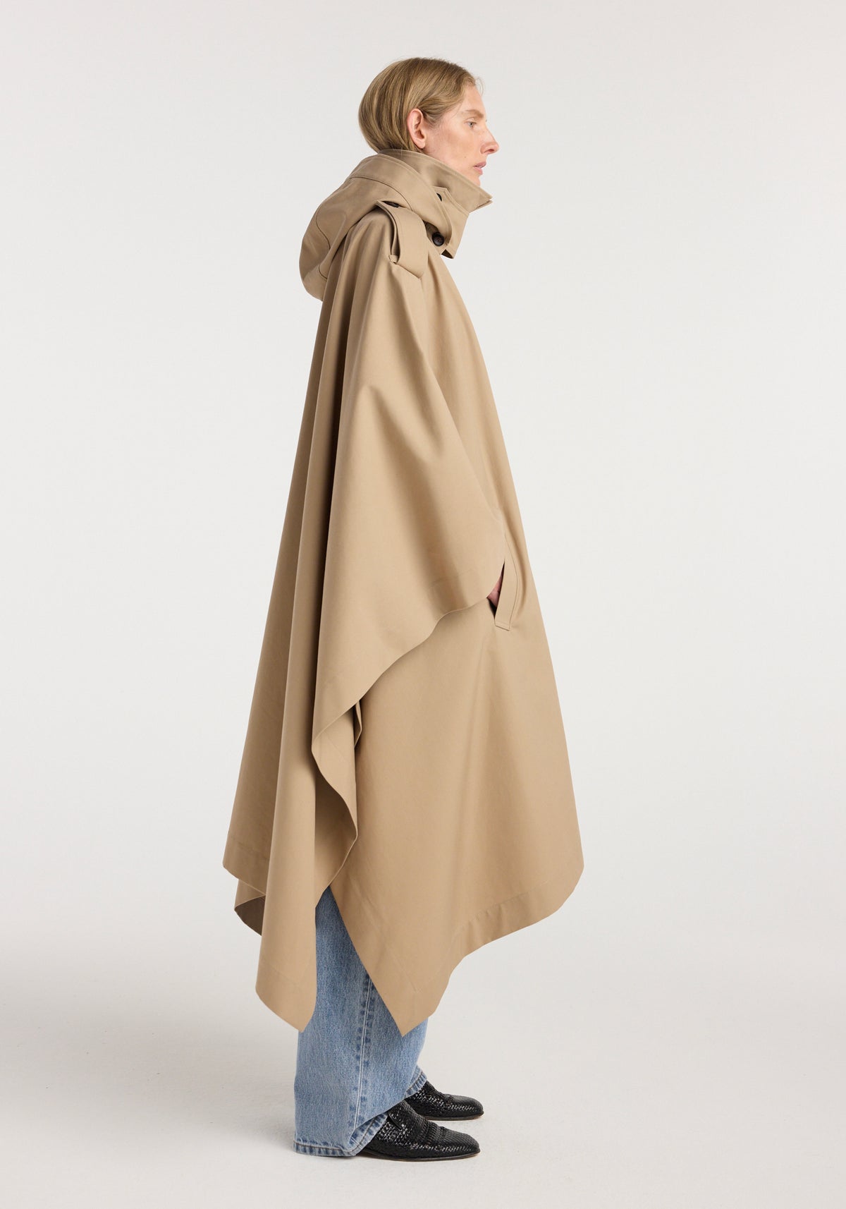Side profile of a person wearing a beige hooded cape trenchcoat | beige over light blue jeans and black shoes against a plain background.