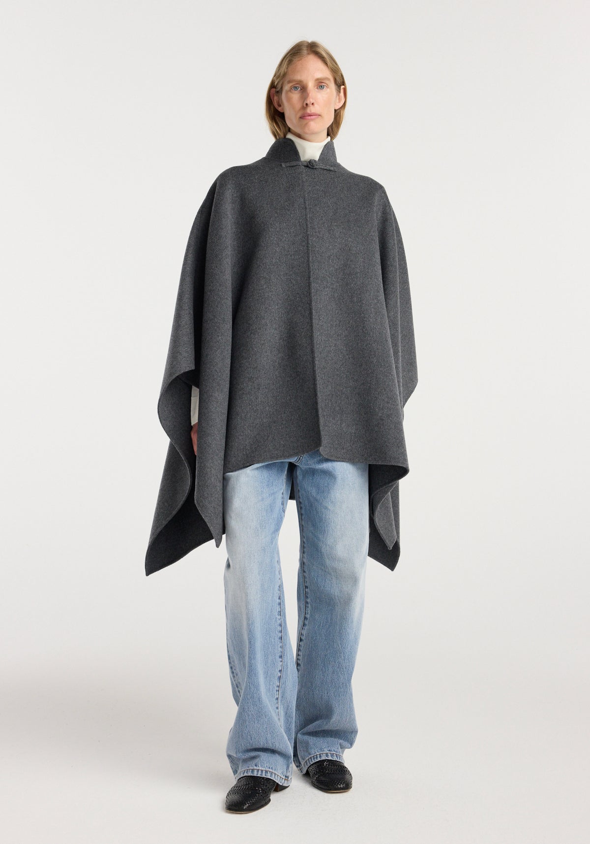 Woman wearing a dark grey melange double-faced wool pankou closure poncho over a white turtleneck and light blue jeans with black shoes against a plain white background, styled in a relaxed fit.