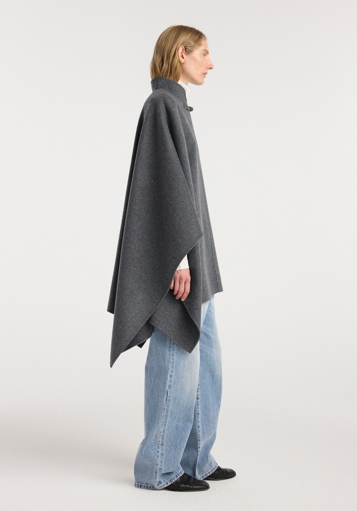 Side view of a person wearing a double-faced wool pankou closure poncho | dark grey melange over a white top with light blue wide-leg jeans and black shoes, standing against a plain background.
