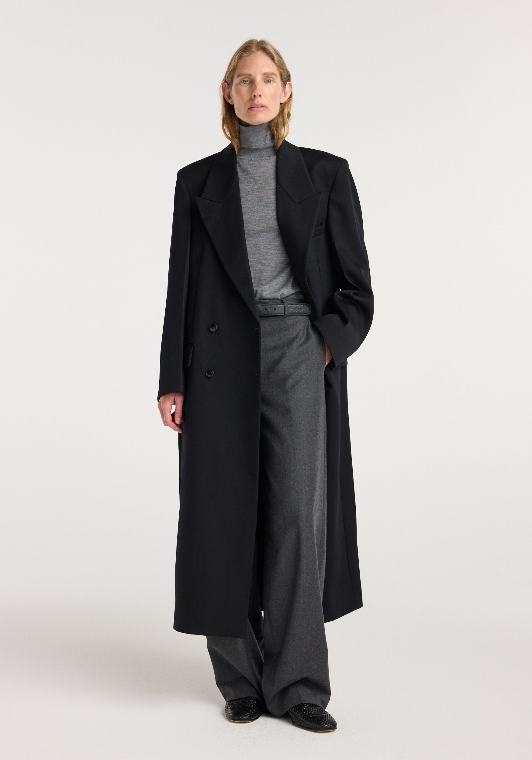 Woman wearing a black double breasted tailored coat | black over a grey turtleneck and wide-leg grey trousers, standing against a plain light background.