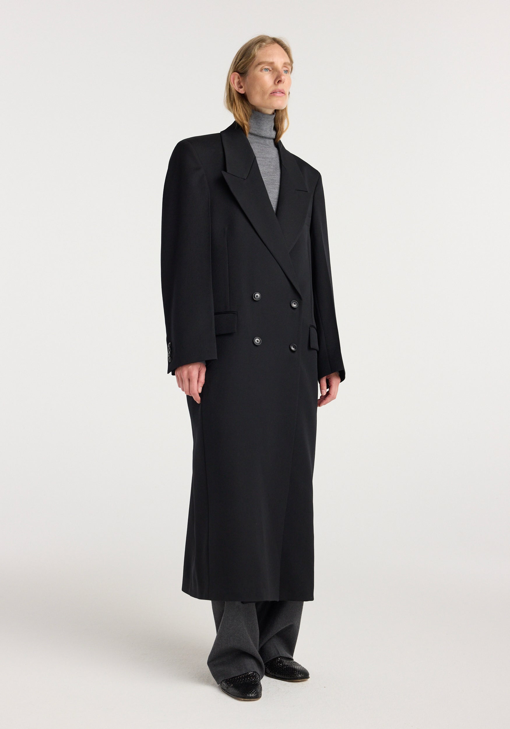 A model wearing a long black double breasted tailored coat | black over a grey turtleneck and grey wide-leg pants, standing against a plain light background.