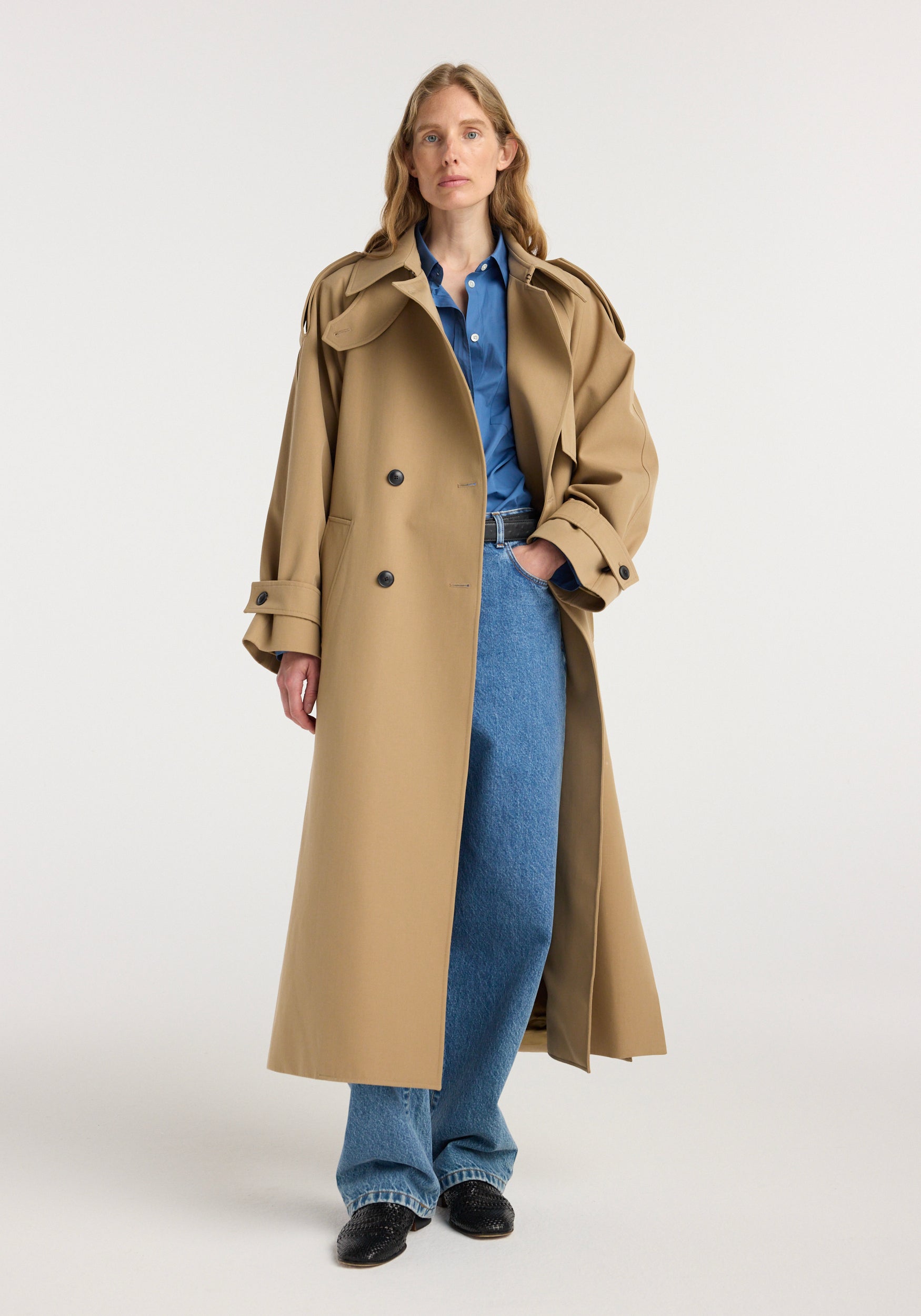Woman wearing a beige classic trench | beige coat over a blue button-up shirt and wide-leg jeans, standing against a plain white background.