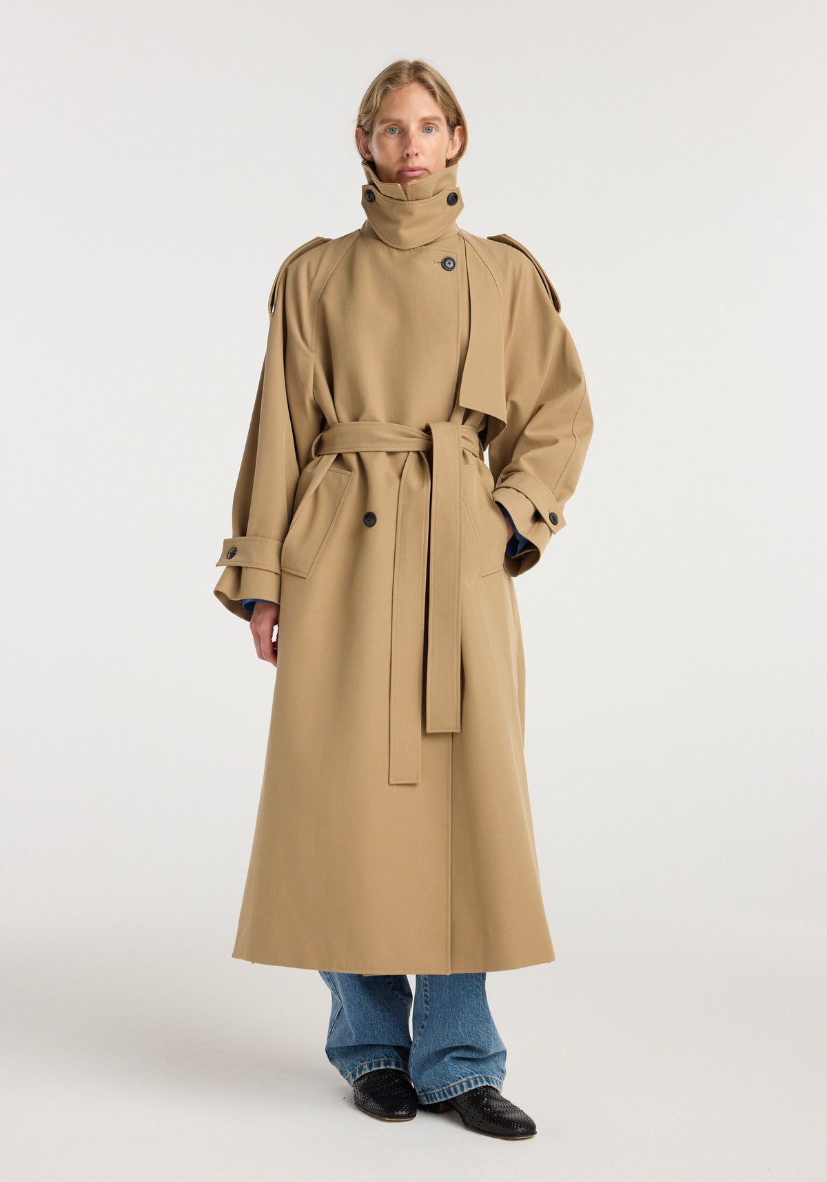 Woman wearing a beige Classic trench | beige coat with high collar, belted waist, and long sleeves, paired with blue jeans and black shoes against a plain background.