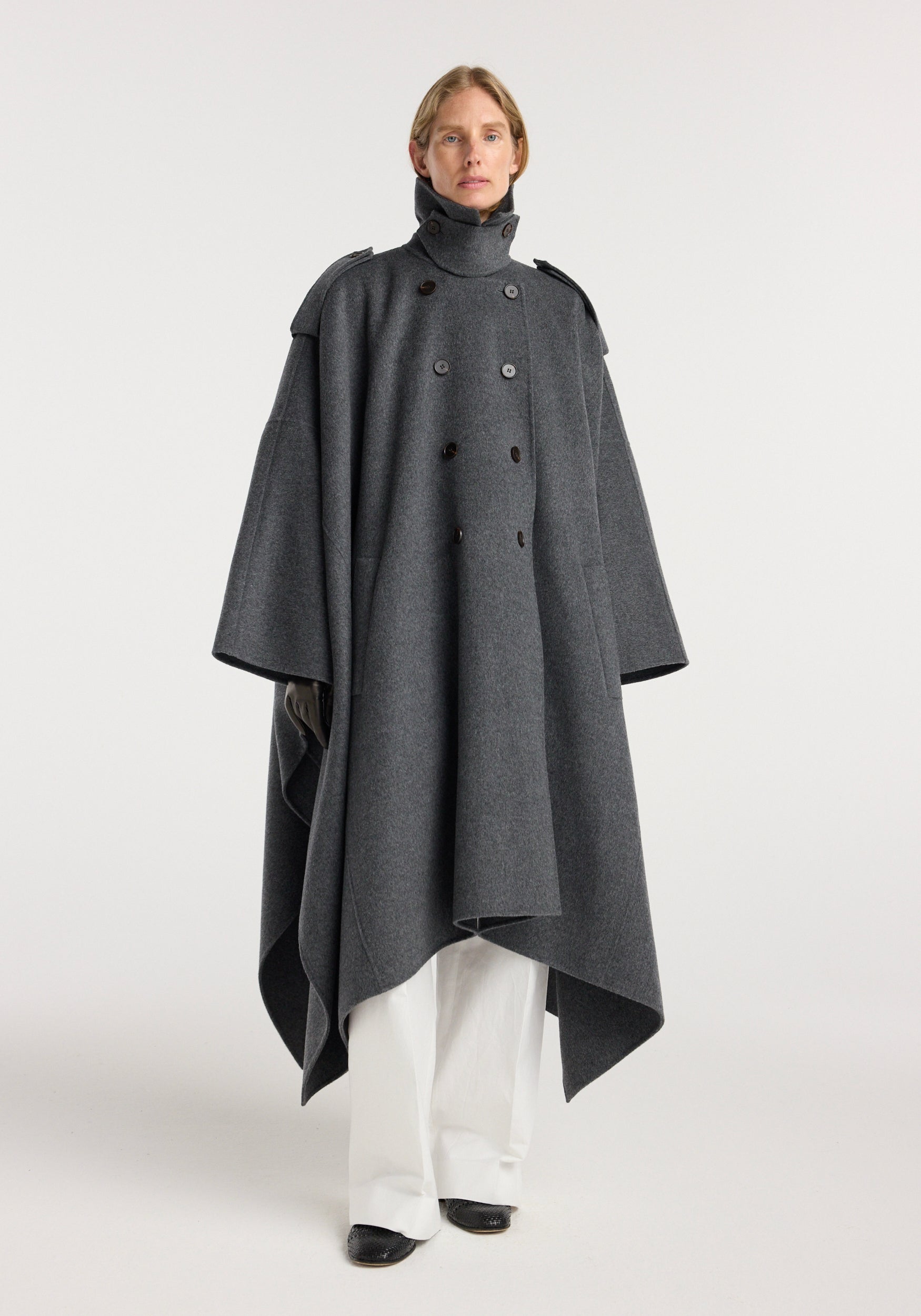A model wearing a double-faced cape trenchcoat | dark grey melange with a high collar and double-breasted button closure, styled with wide white pants and black shoes.