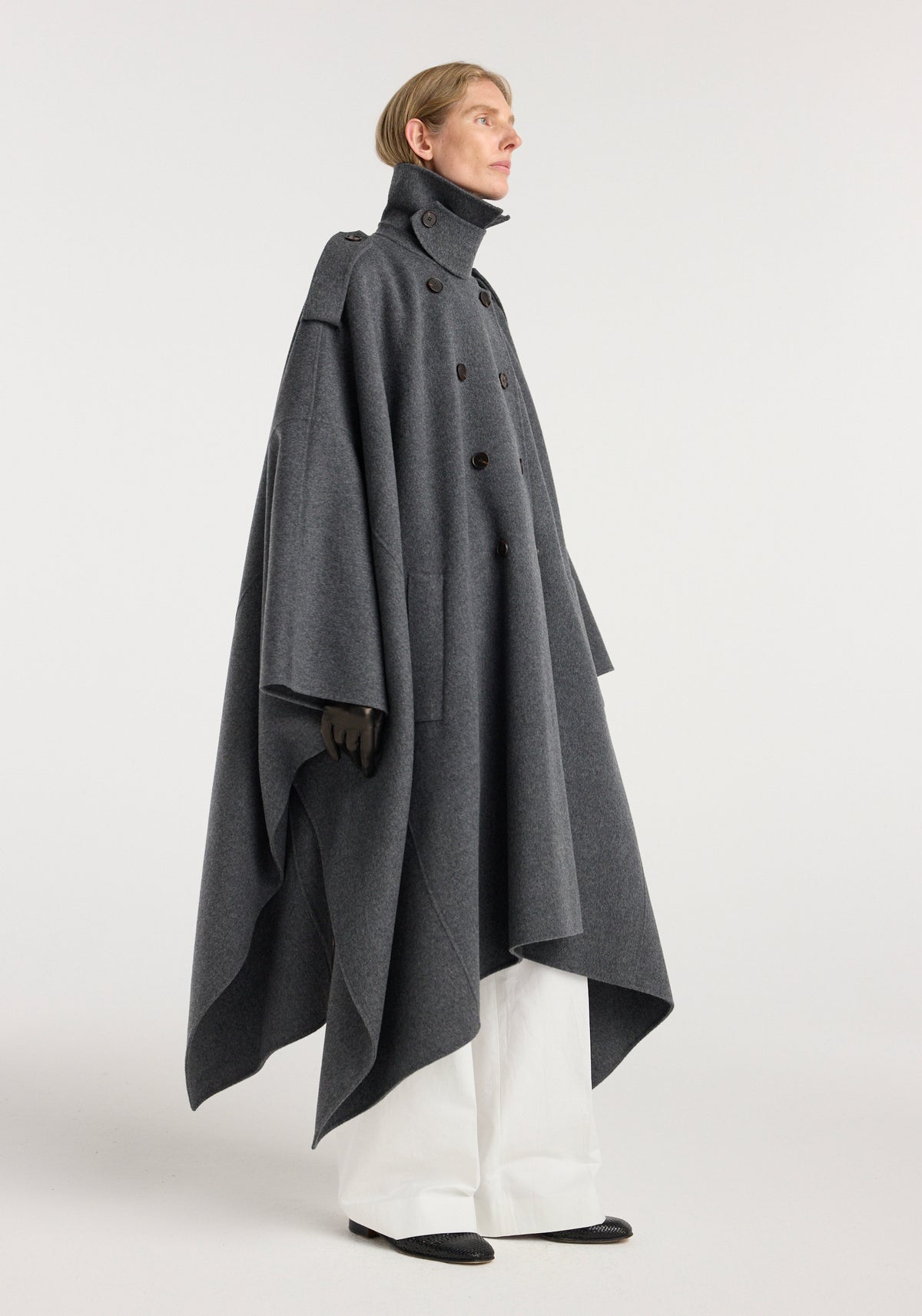 Model wearing a dark grey melange double-faced cape trenchcoat with a high collar, button closure, and wide draped silhouette over white pants and black gloves, standing in profile.