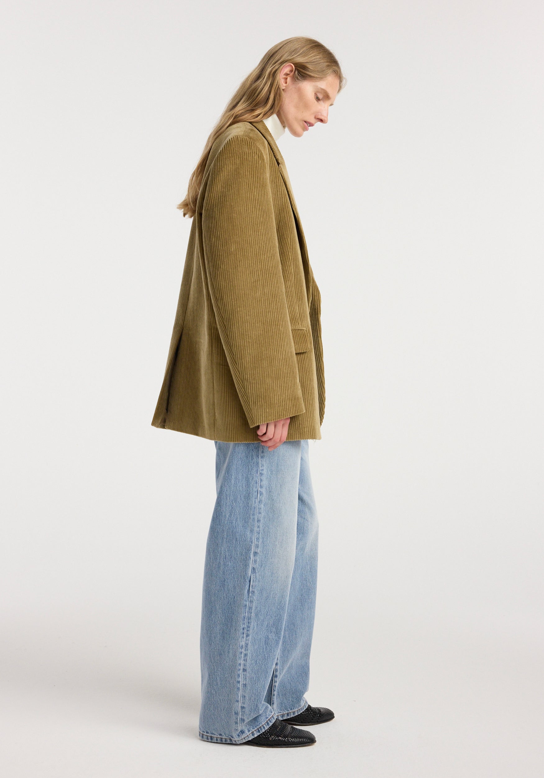 Side view of a woman wearing an oversized corduroy blazer jacket | beige paired with light blue wide-leg jeans and black pointed-toe shoes against a plain background.