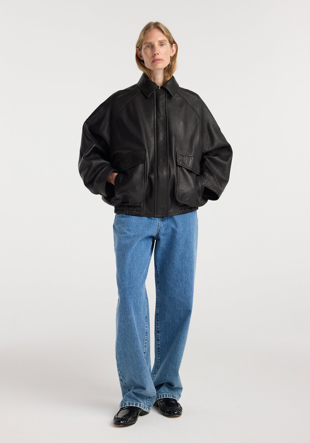 A model wearing a black Grain leather aviator jacket | black with a front zipper, large pockets, and curved sleeves paired with loose blue jeans and black shoes.