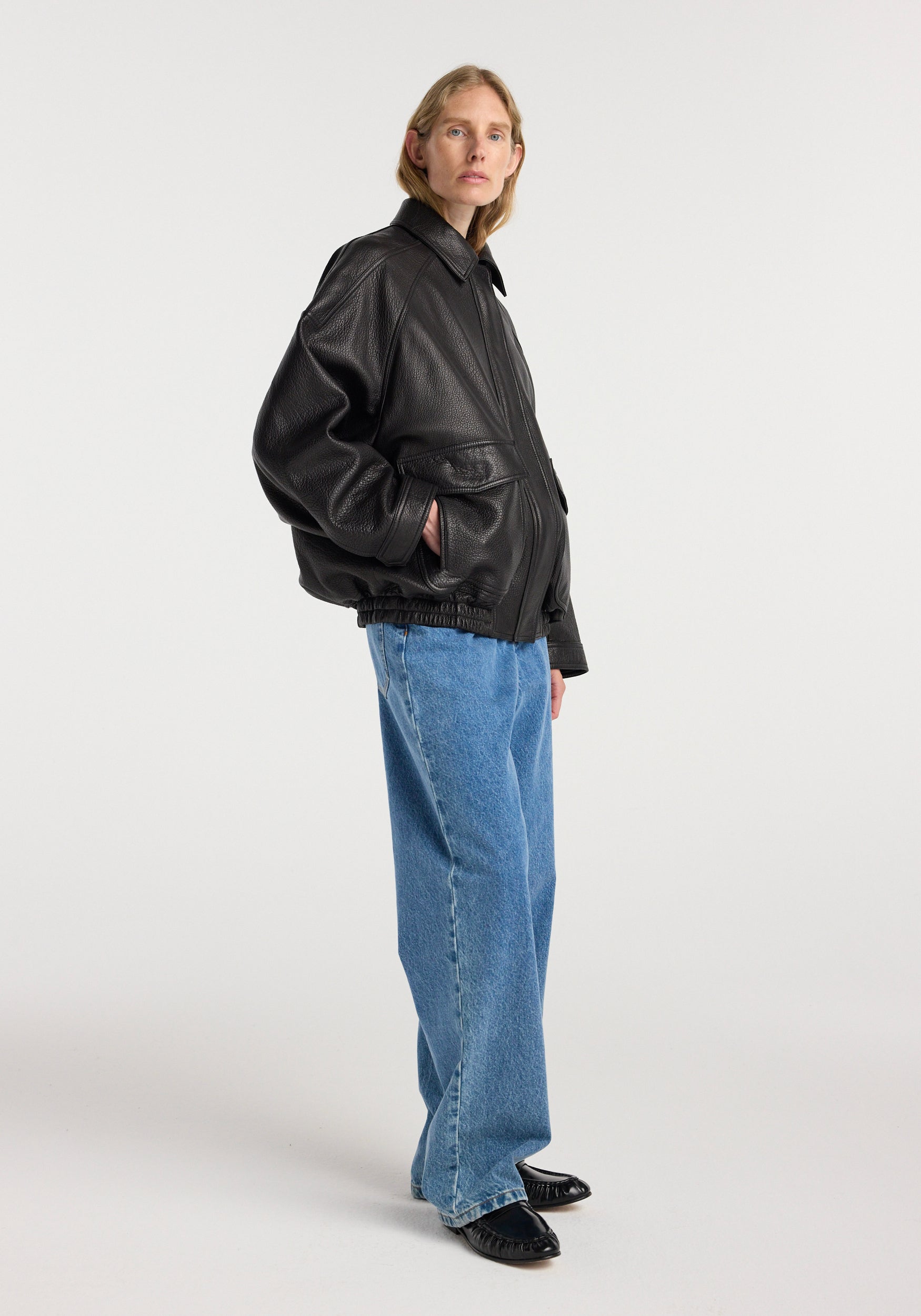 Woman wearing a black grain leather aviator jacket | black with side pockets, paired with loose blue jeans and black shoes, standing against a plain white background.