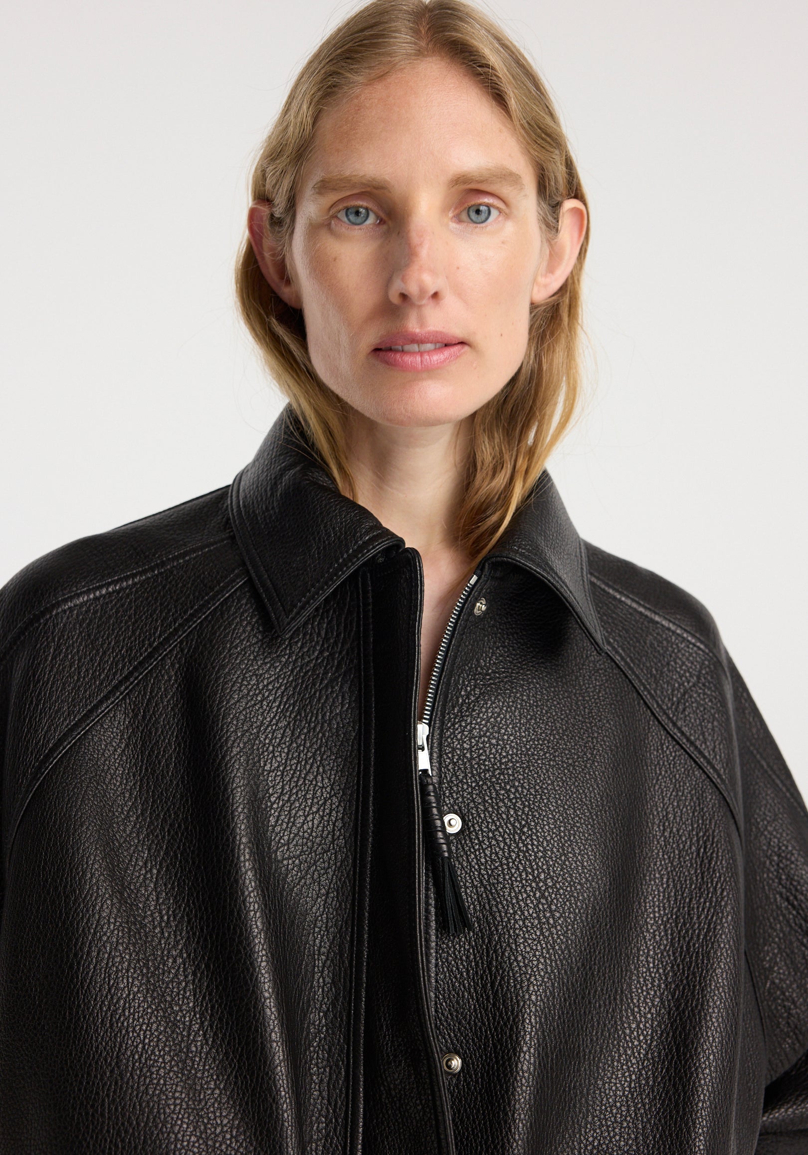 Close-up of a woman wearing a black grain leather aviator jacket | black with a textured finish, wide collar, and front zipper against a plain white background.