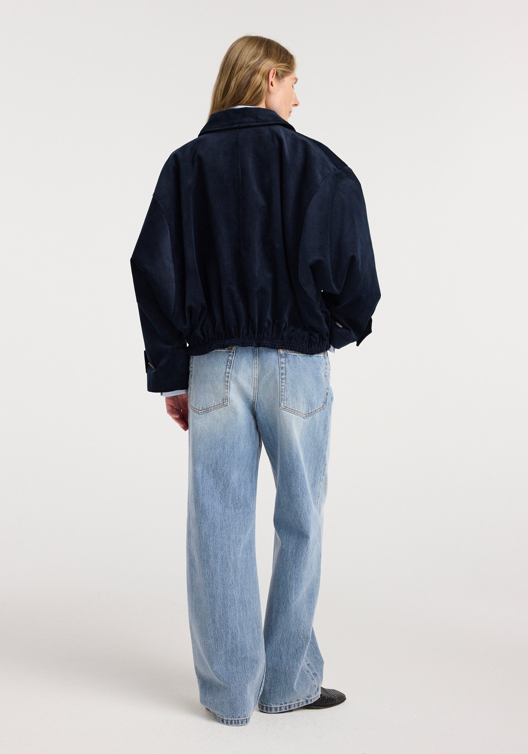 A person seen from the back wearing a dark navy corduroy blouson jacket | night with elastic gathering at the waist paired with light blue straight-leg jeans against a plain white background.