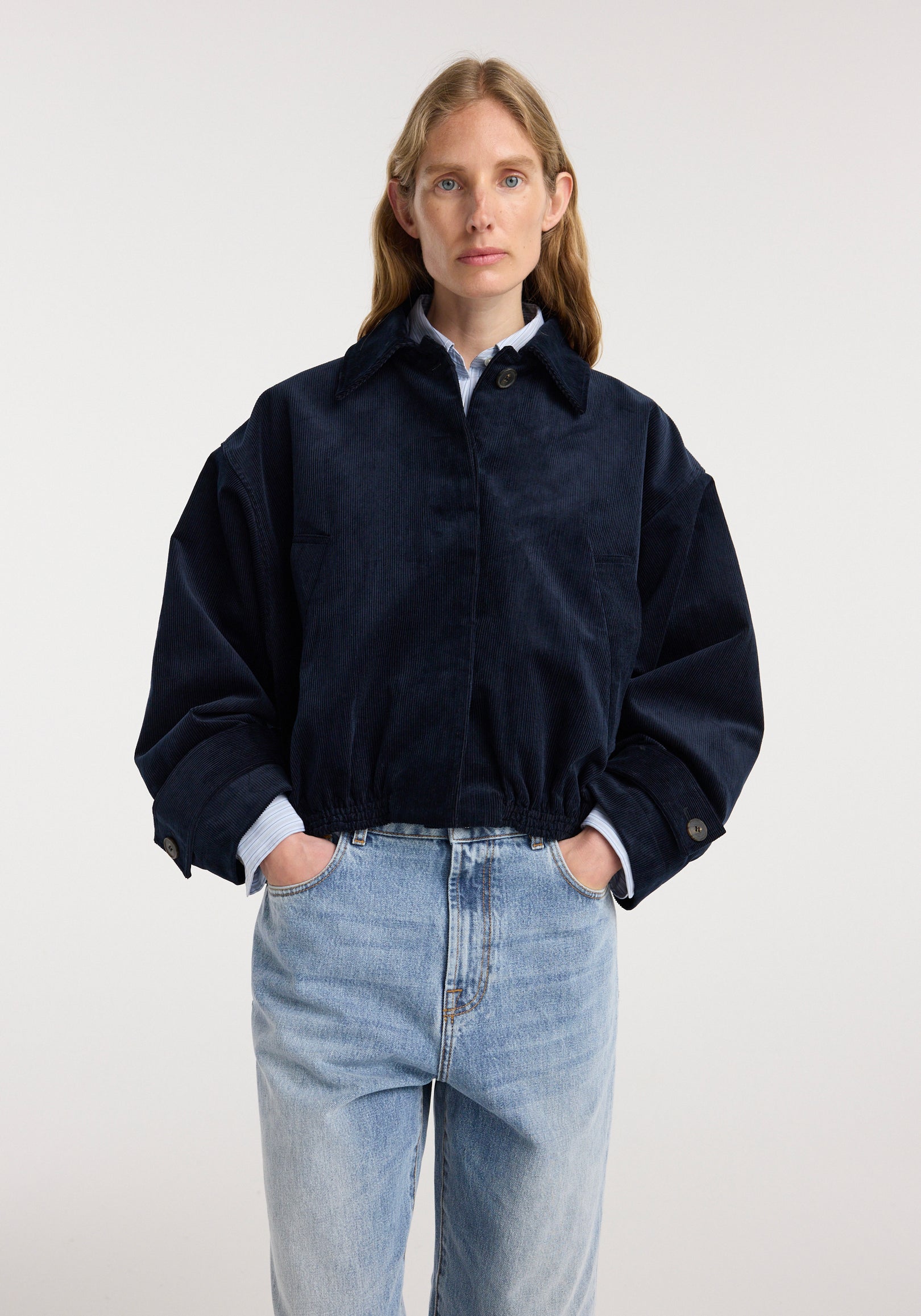 Woman wearing a dark navy Corduroy blouson jacket | night with button closure and elastic hem, paired with light blue jeans, standing against a plain background.