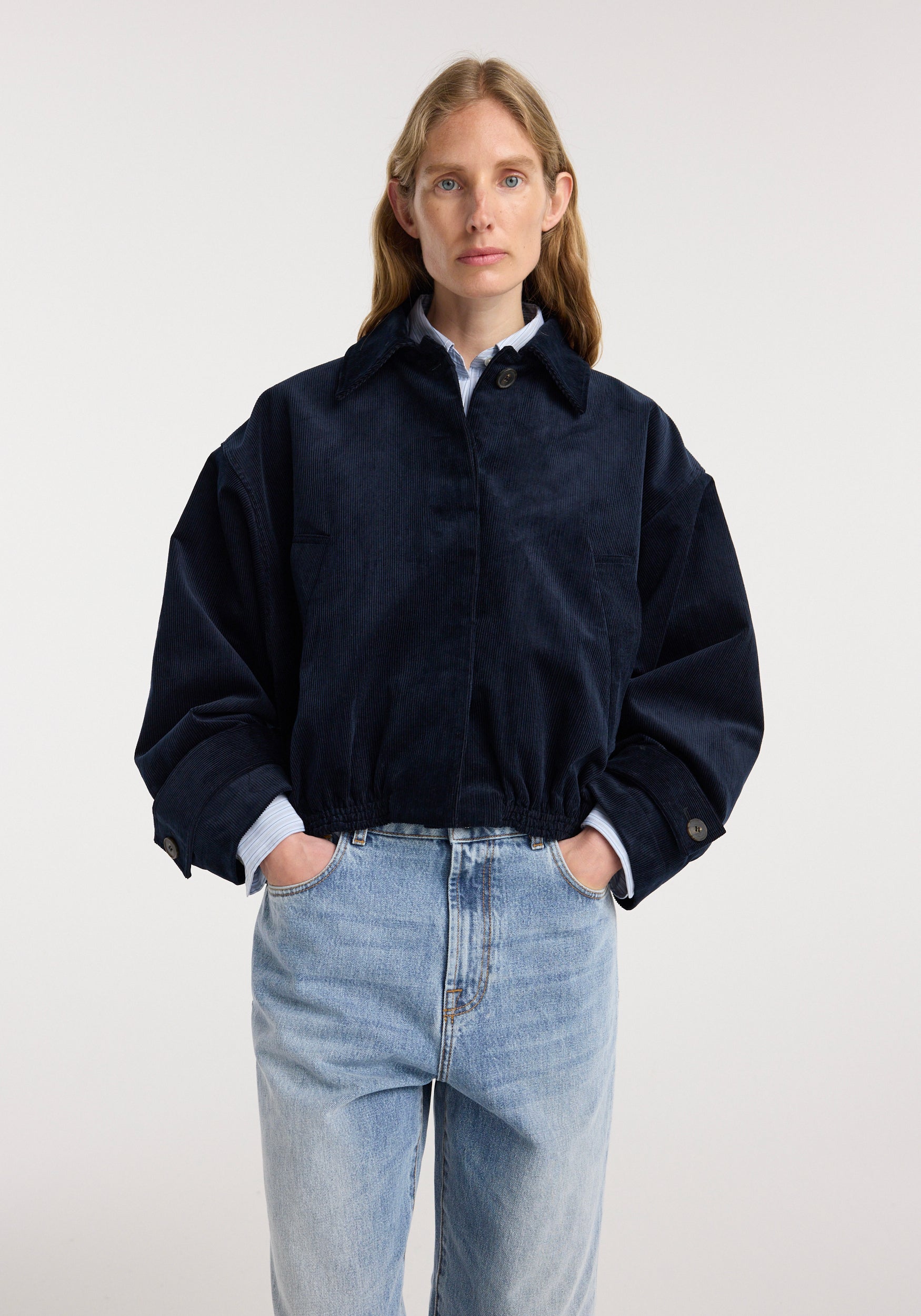 Woman wearing a dark navy Corduroy blouson jacket | night with button closure and elastic hem, paired with light blue jeans, standing against a plain background.