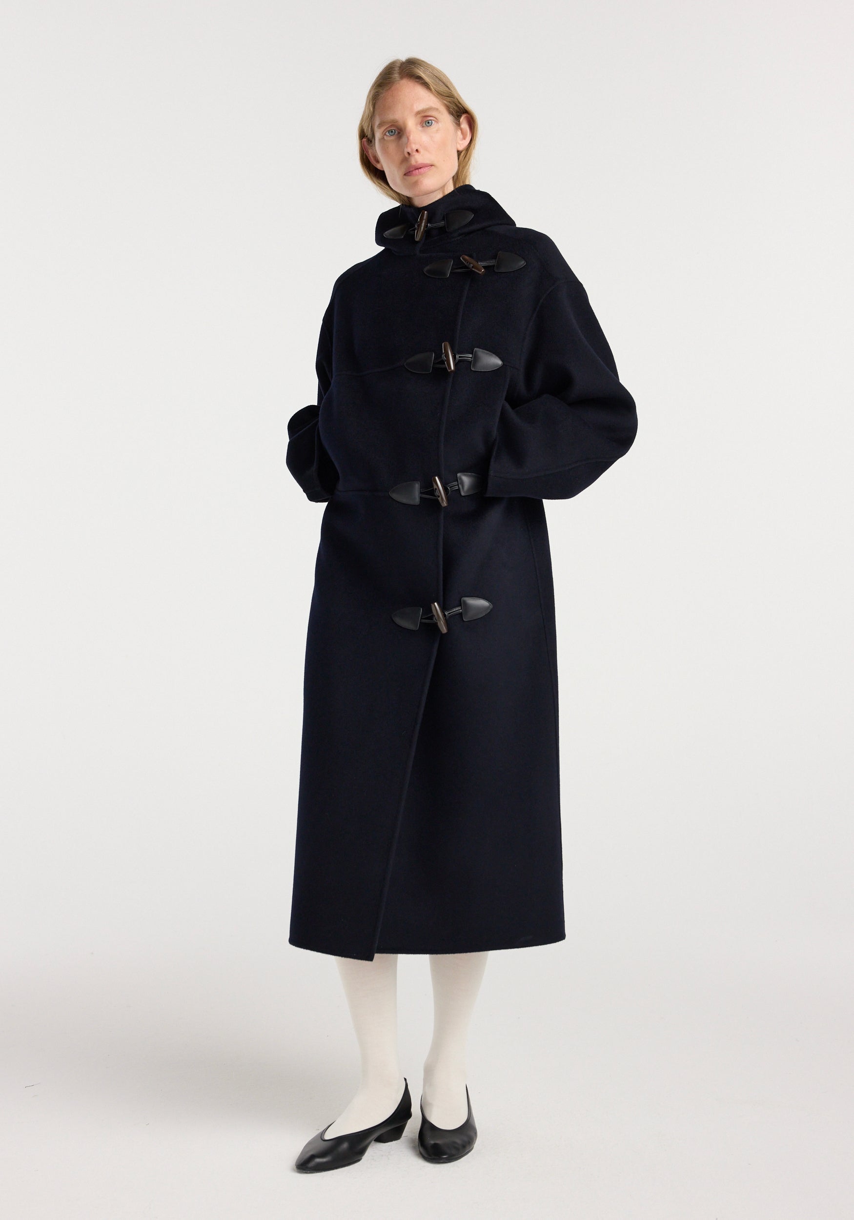 A model wearing a long navy double-faced wool duffel coat with toggle closures, a hood, and side pockets, paired with white tights and black flats.