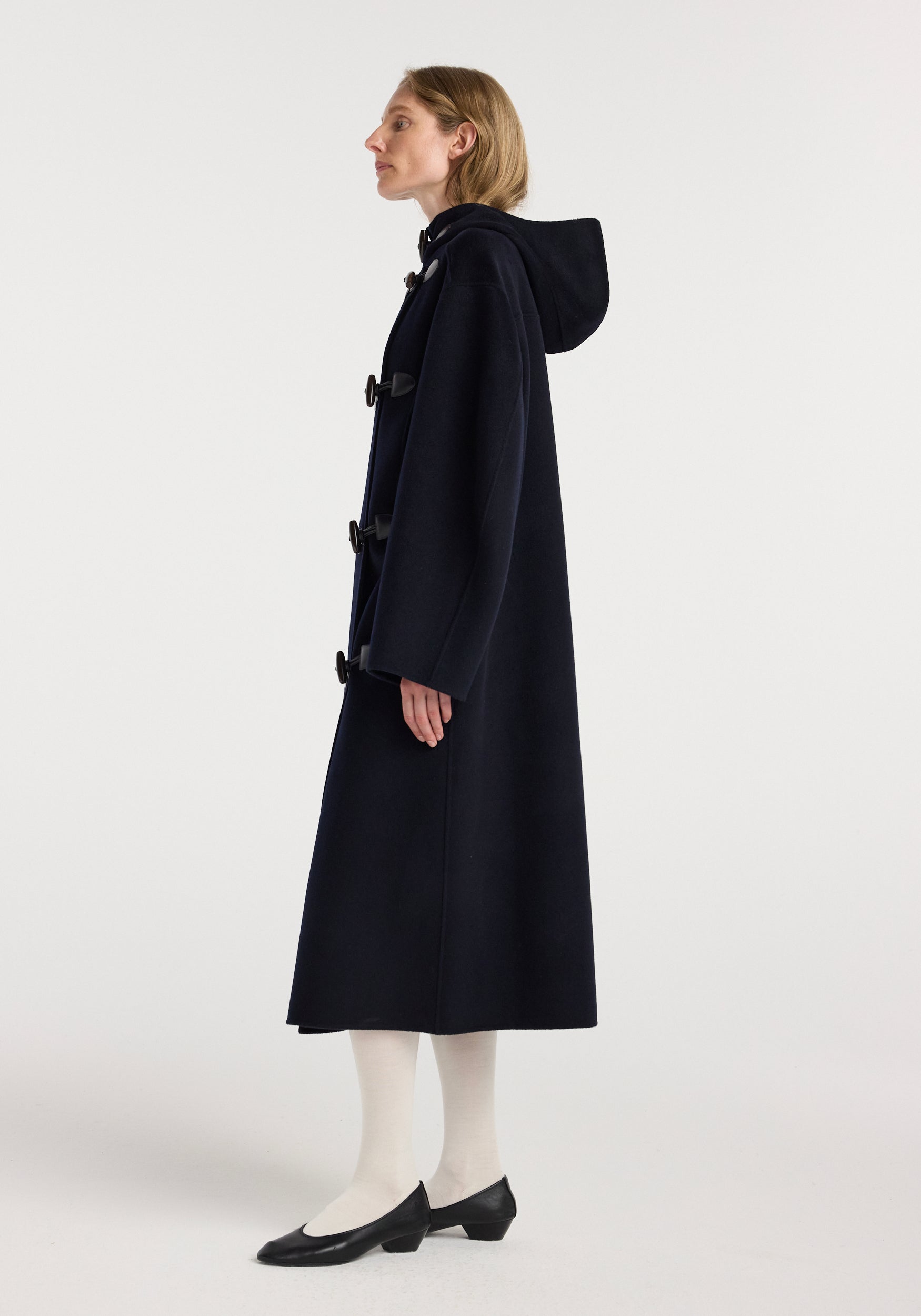 Woman wearing a long, navy double-faced wool duffel coat with toggle closures and a hood, paired with white tights and black low-heeled shoes in a side profile pose.