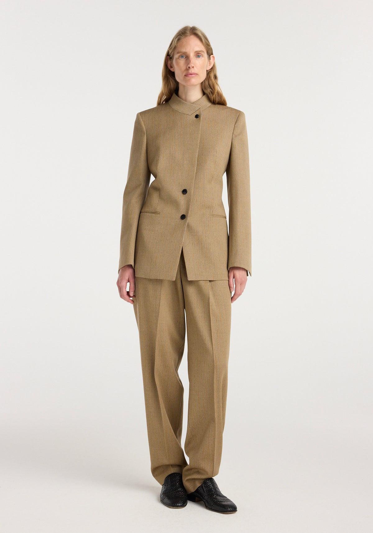 A woman wearing a tan striped suit consisting of a tailored overlap lapel blazer | heritage stripe and matching wide-leg trousers with black shoes, standing against a plain white background.