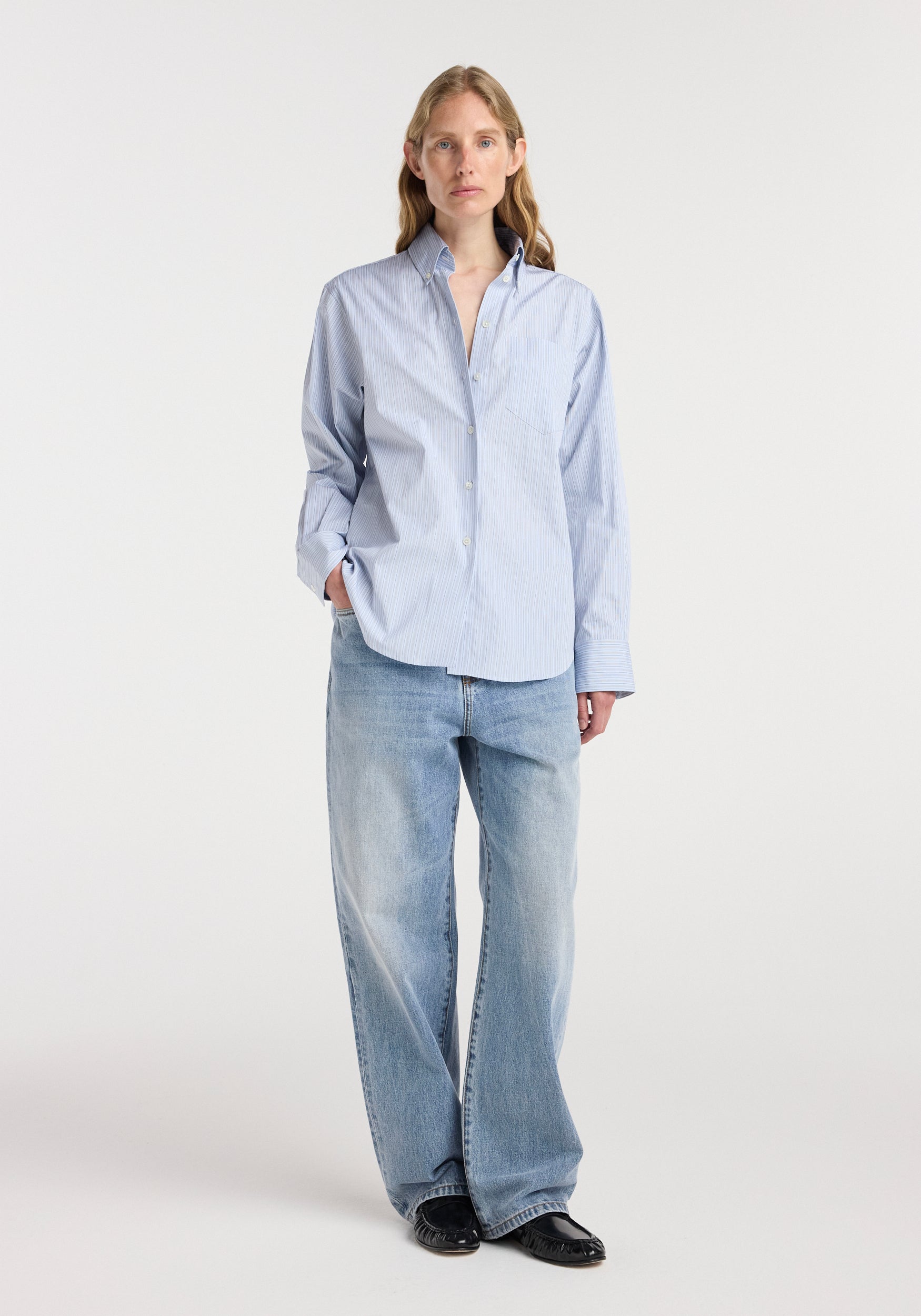Woman wearing a light blue and white Oxford striped shirt | blue stripe with long sleeves, tucked casually into light wash wide-leg jeans and black shoes, standing against a white background.