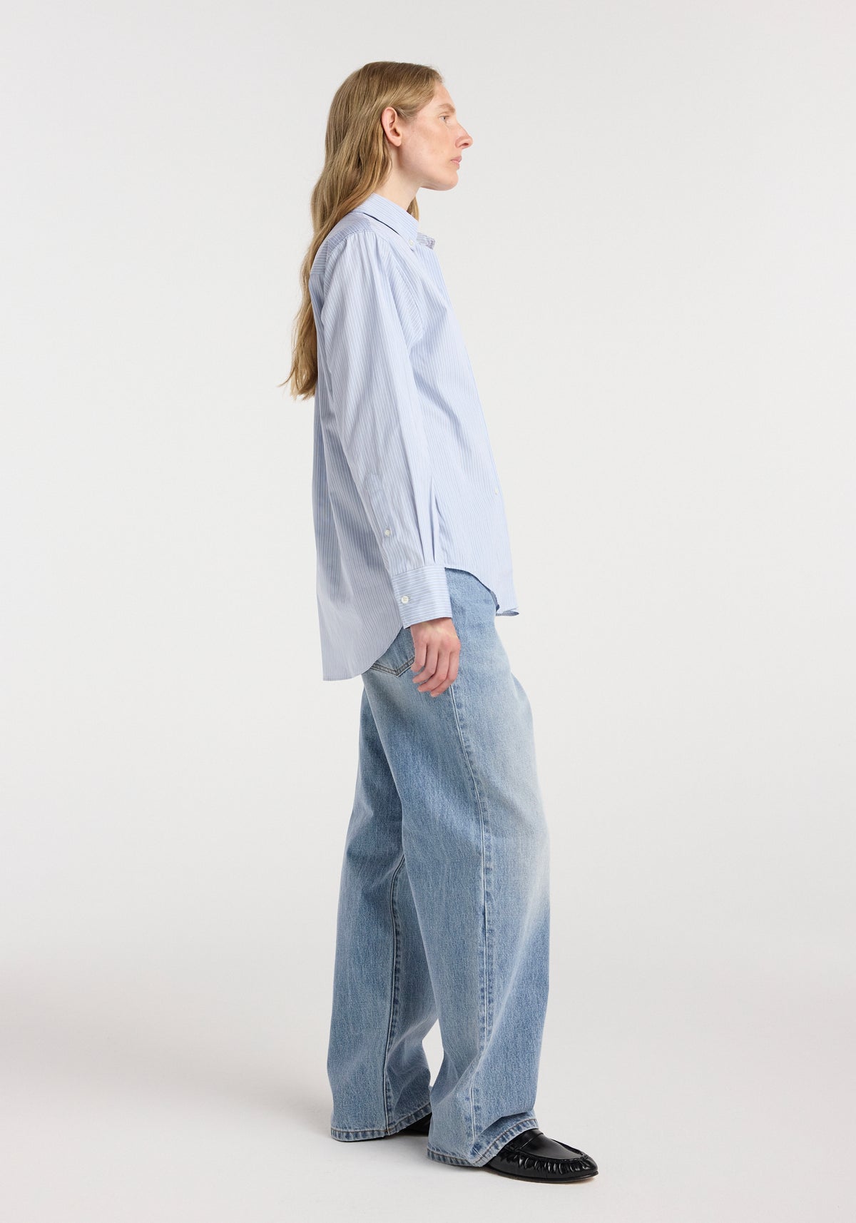 Side view of a woman wearing an Oxford striped shirt | blue stripe paired with light blue wide-leg jeans and black shoes against a plain white background.