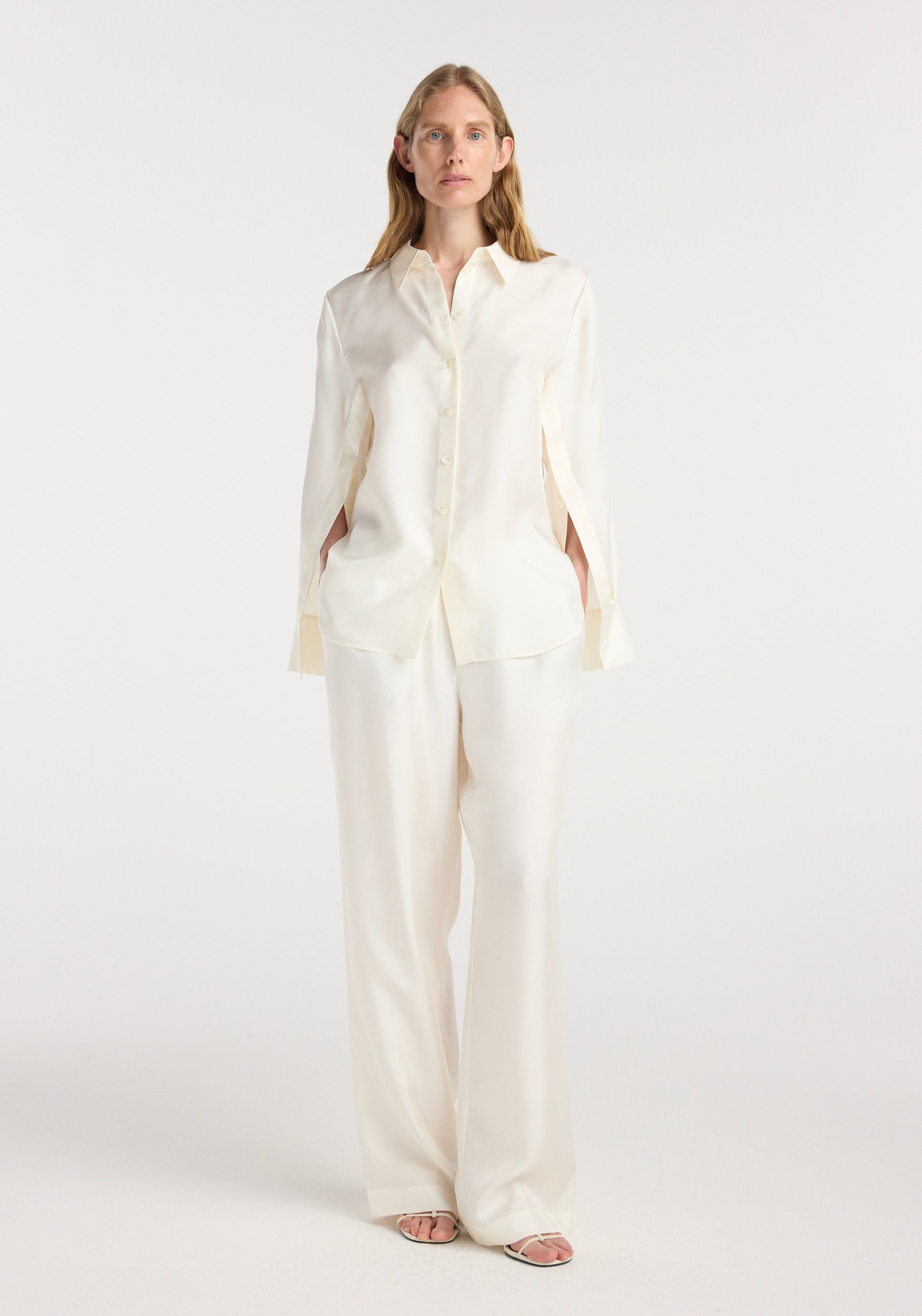 Woman wearing a cream Silk open-sleeve detailed shirt | cream with button front and wide-leg cream pants standing against a white background.