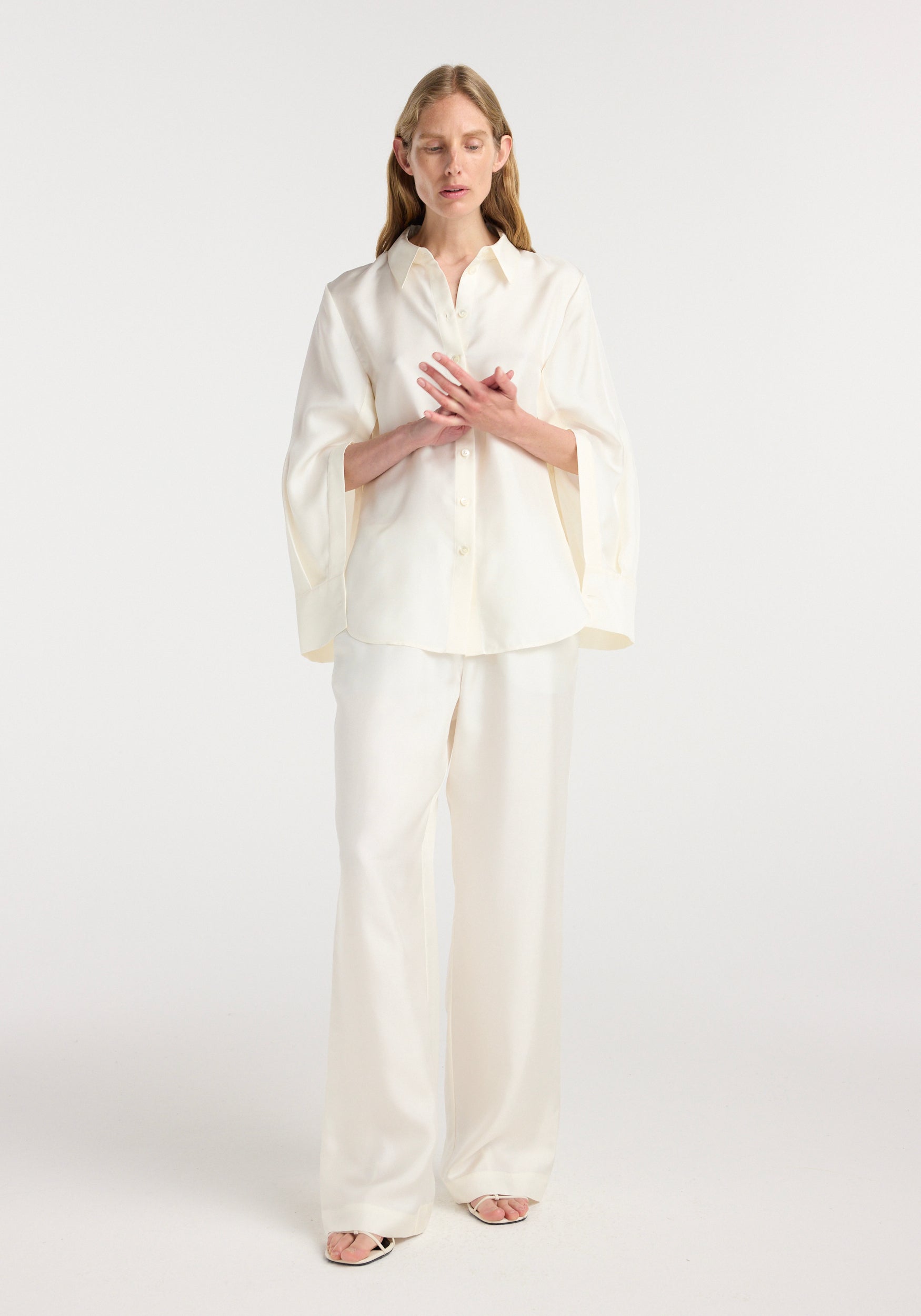Woman wearing a cream silk open-sleeve detailed shirt | cream paired with matching wide-leg pants, standing with hands crossed at the chest against a plain background.