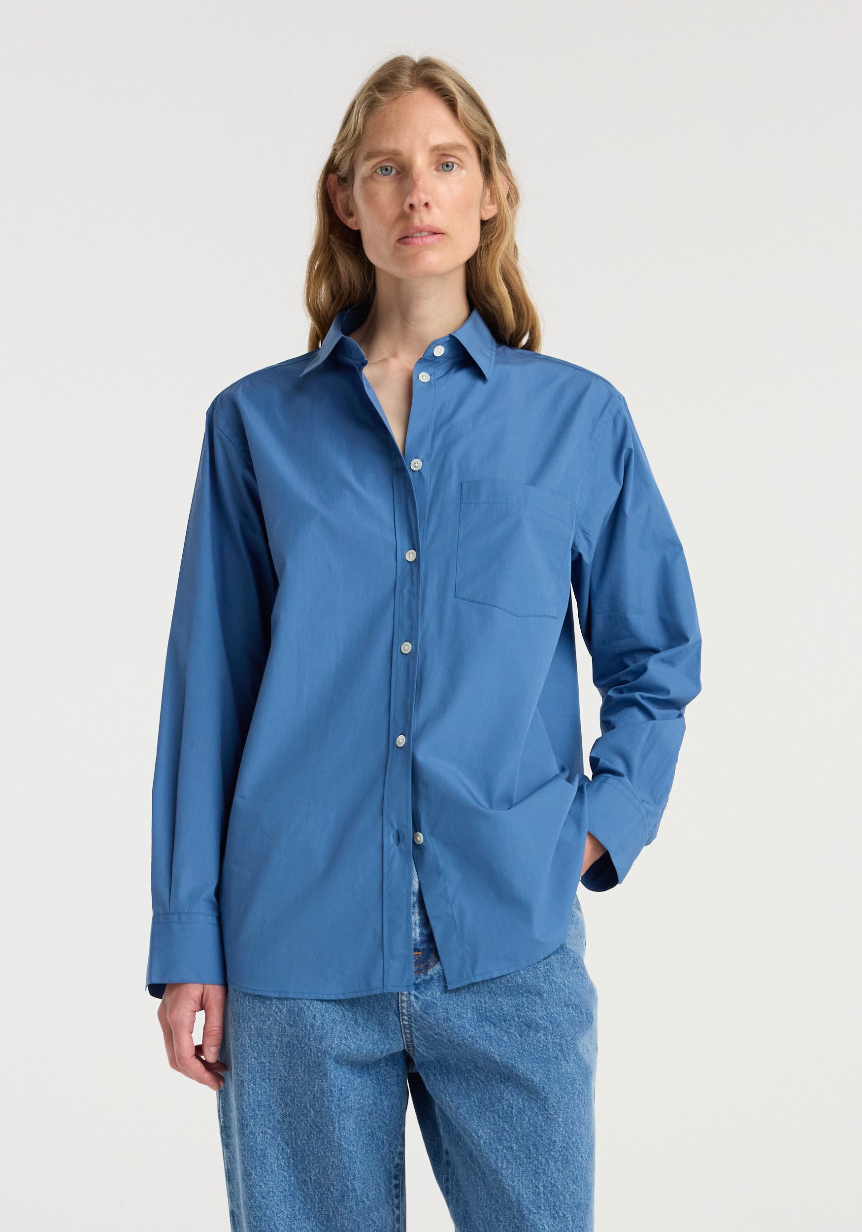 Woman wearing a classic fit shirt | pacific blue, styled with long sleeves and a chest pocket, paired with light blue jeans against a plain background.