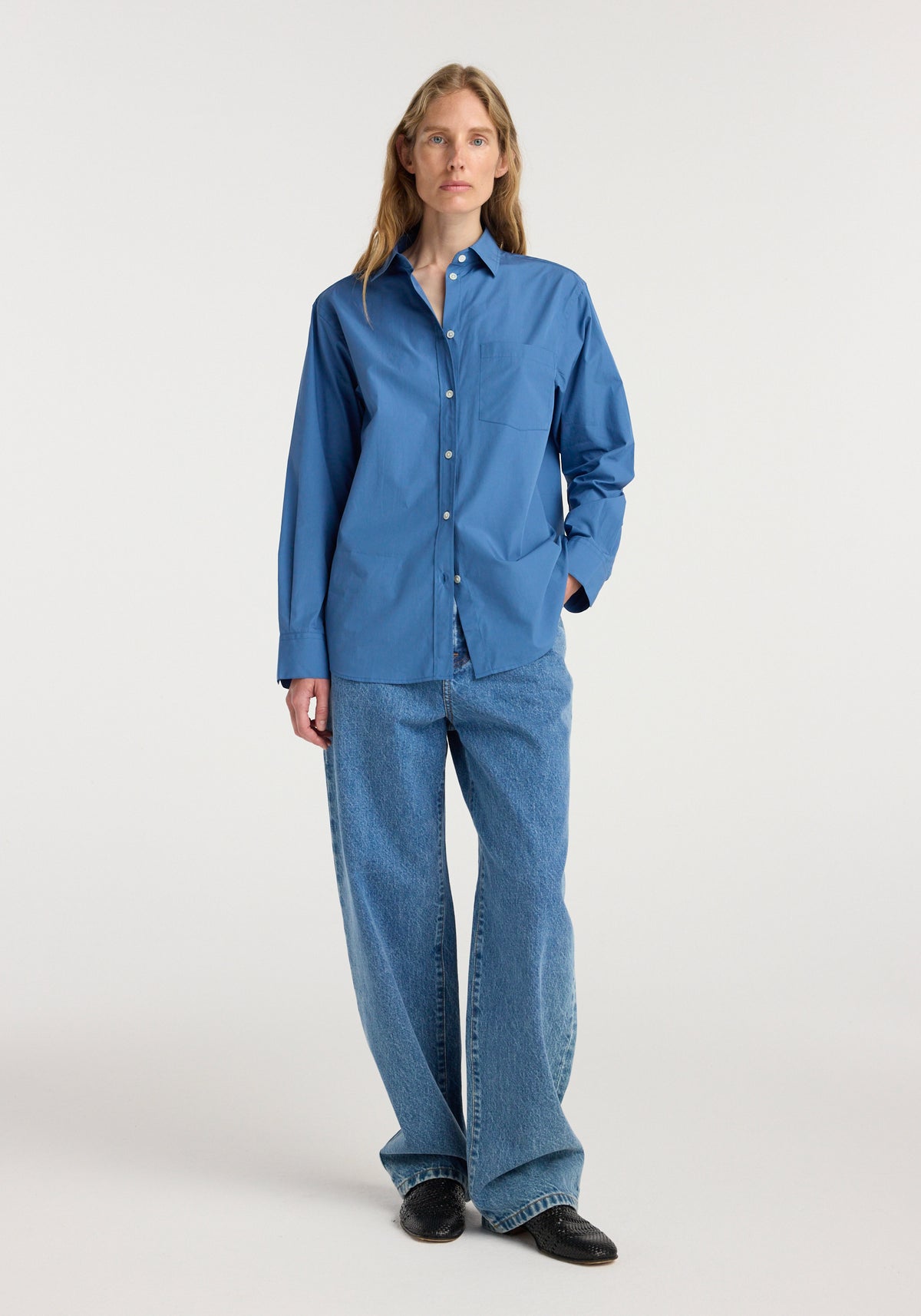 A woman wearing a Classic fit shirt | pacific blue paired with loose-fitting blue jeans and black shoes, standing against a plain white background.