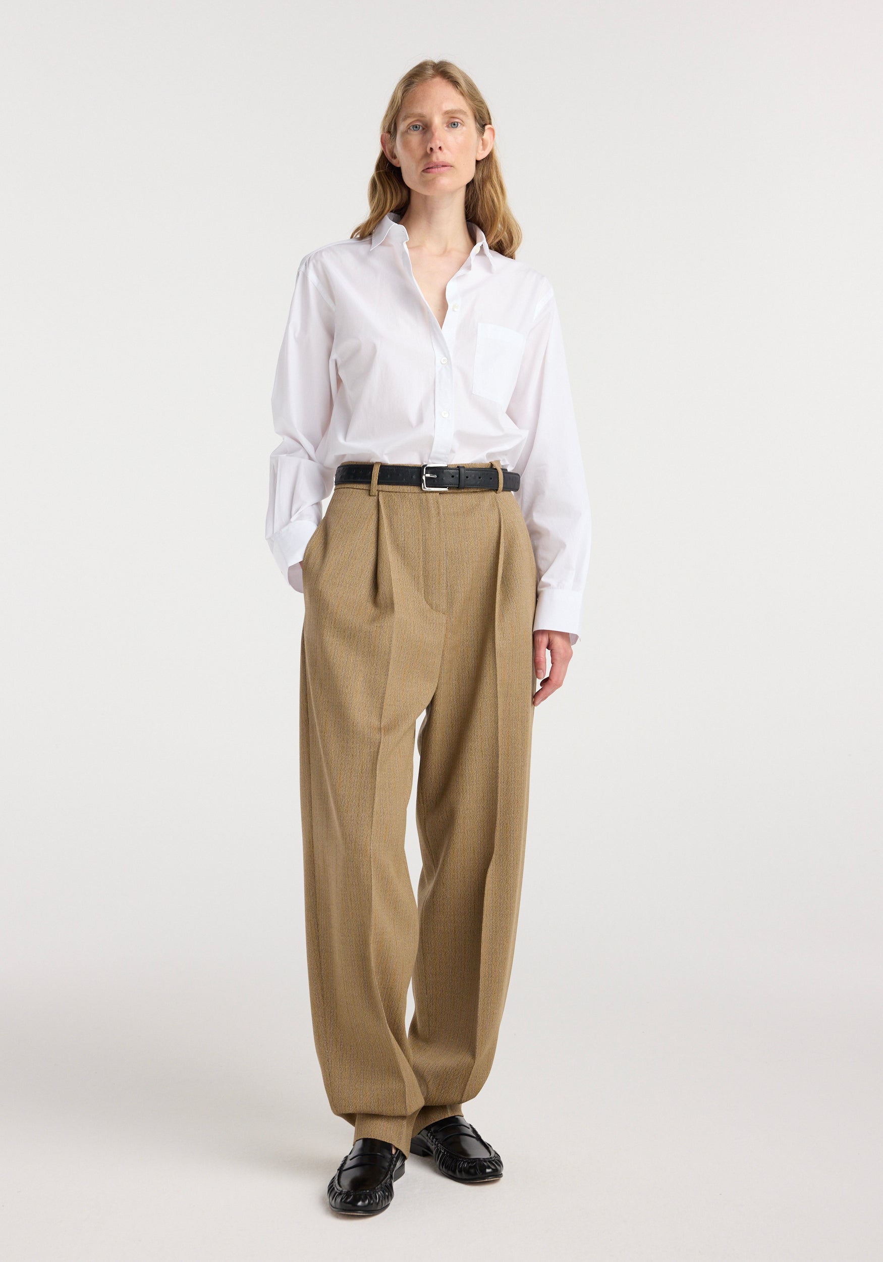 Woman wearing a Classic fit shirt | optic white paired with high-waisted beige pleated trousers, black belt, and black shoes, standing against a plain background.