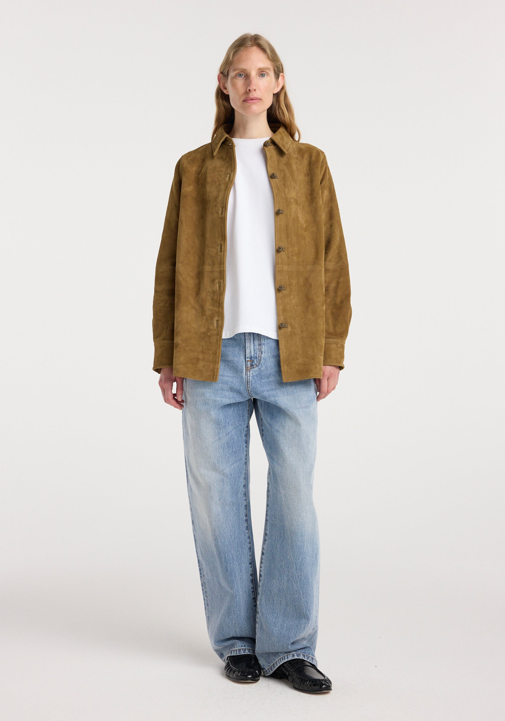 A woman wearing a sage suede pankou overshirt over a white top paired with light blue wide-leg jeans and black shoes, standing against a plain background.