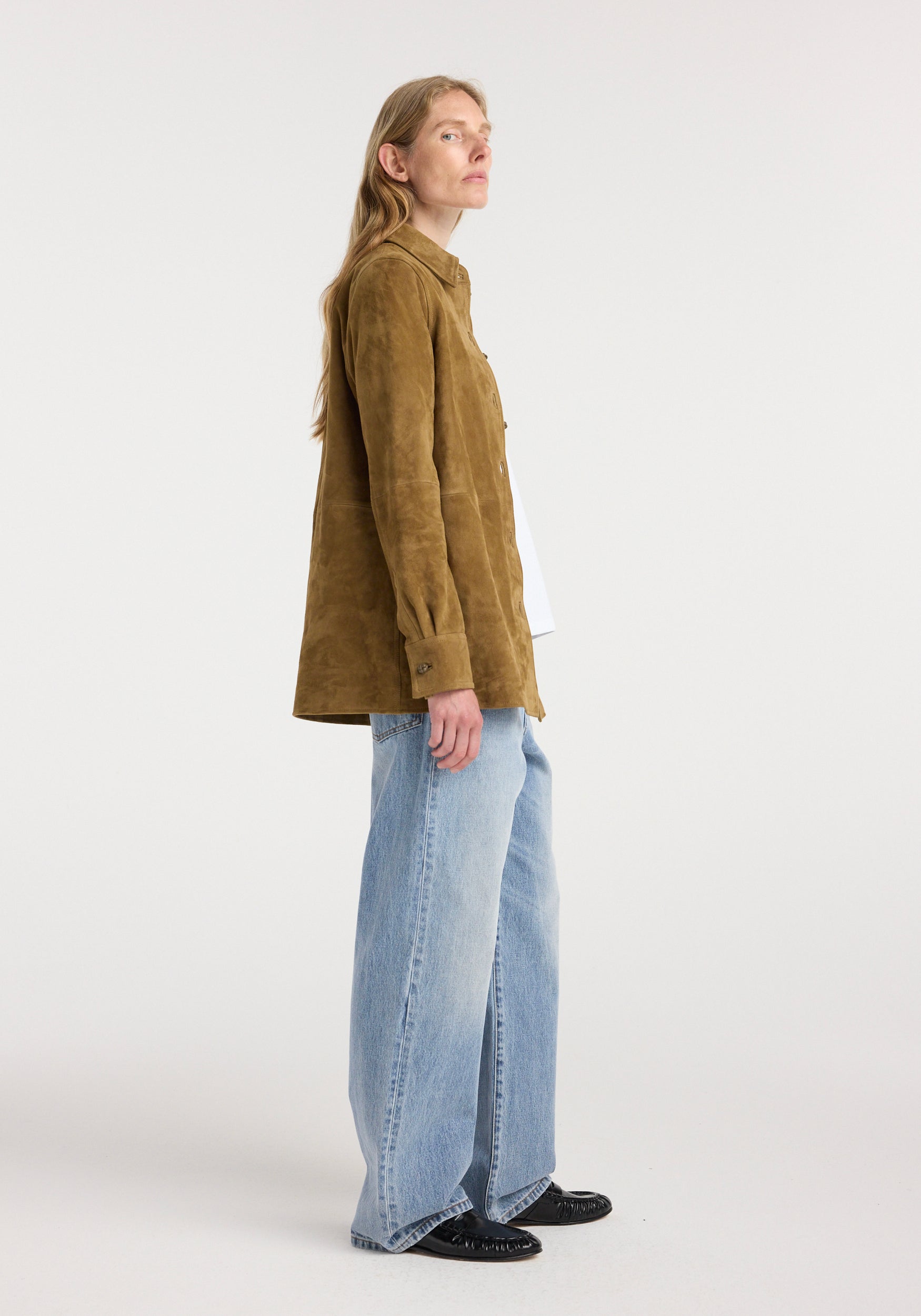 Side view of a woman wearing a brown suede pankou overshirt | sage with buttoned cuffs, paired with light blue wide-leg jeans and black shoes against a white background.
