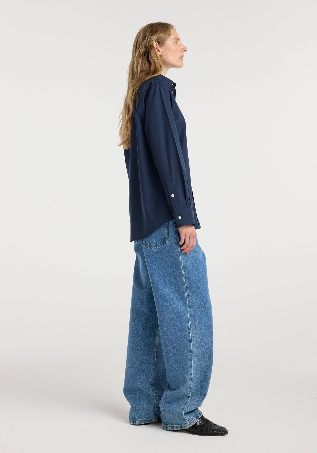 Woman wearing a Classic fit shirt | navy with long sleeves, paired with loose-fitting blue jeans and black shoes, standing in profile against a plain background.