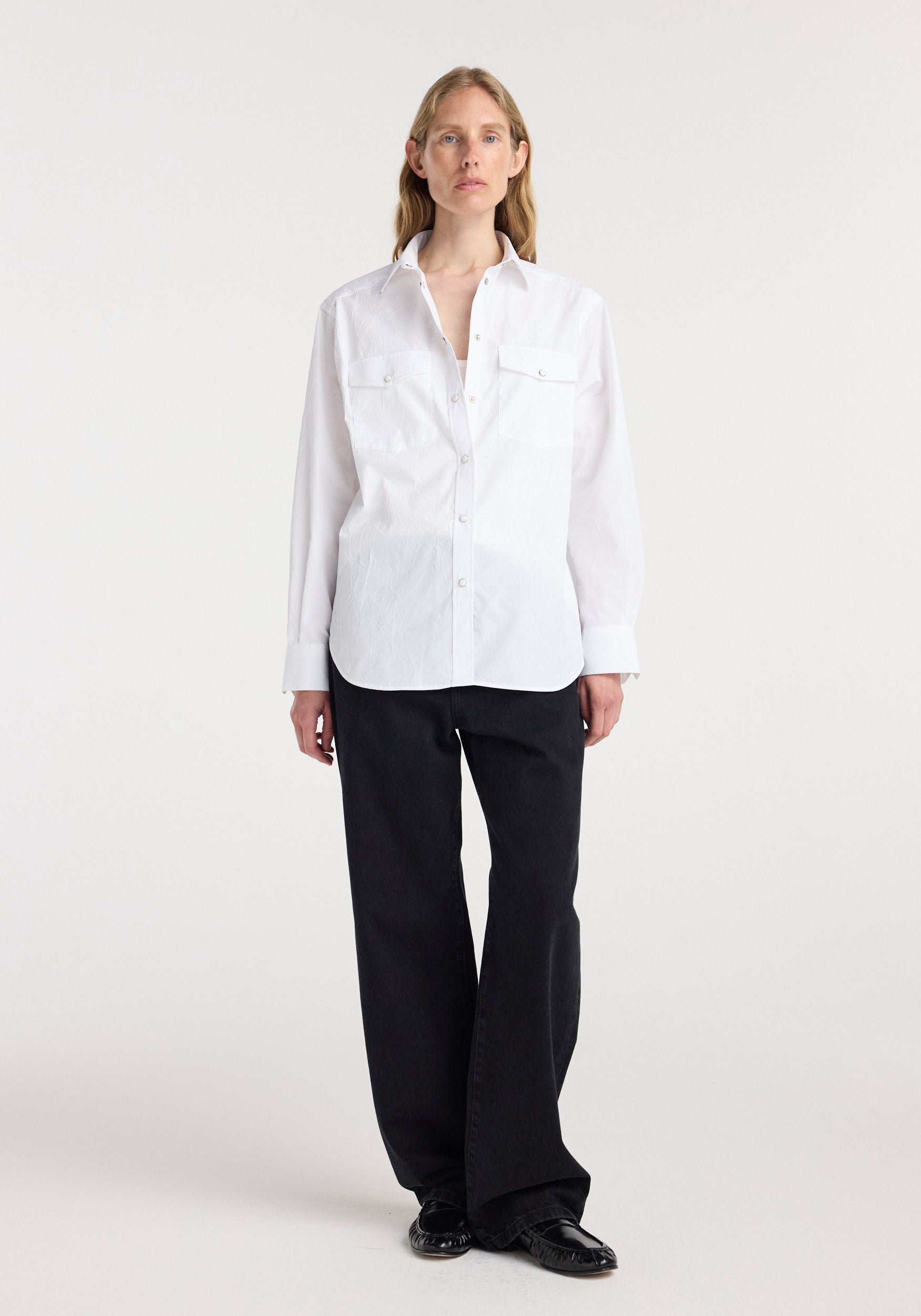 Woman wearing a white Western shirt | optic white with snap buttons and two chest pockets, paired with black wide-leg pants and black shoes, standing against a plain background.