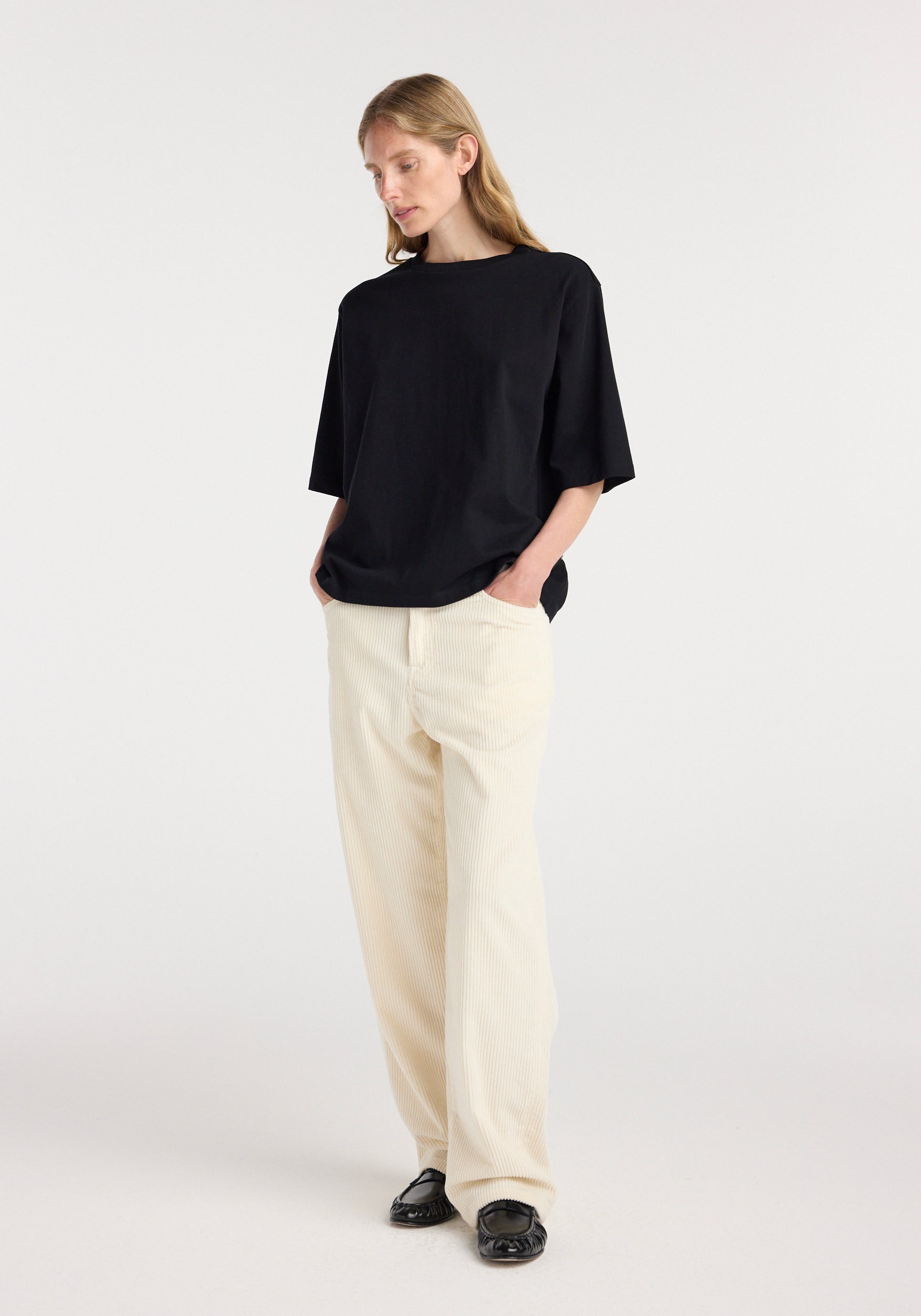 Woman wearing an oversized cotton t-shirt | black paired with loose cream-colored pants and black shoes, standing with hands in pockets against a plain background.