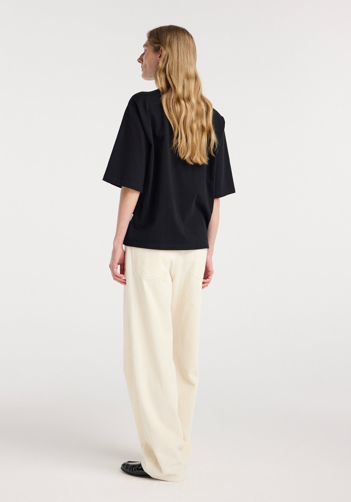 Woman seen from the back wearing an oversized cotton t-shirt | black with loose-fitting light-colored pants and black shoes against a plain background.