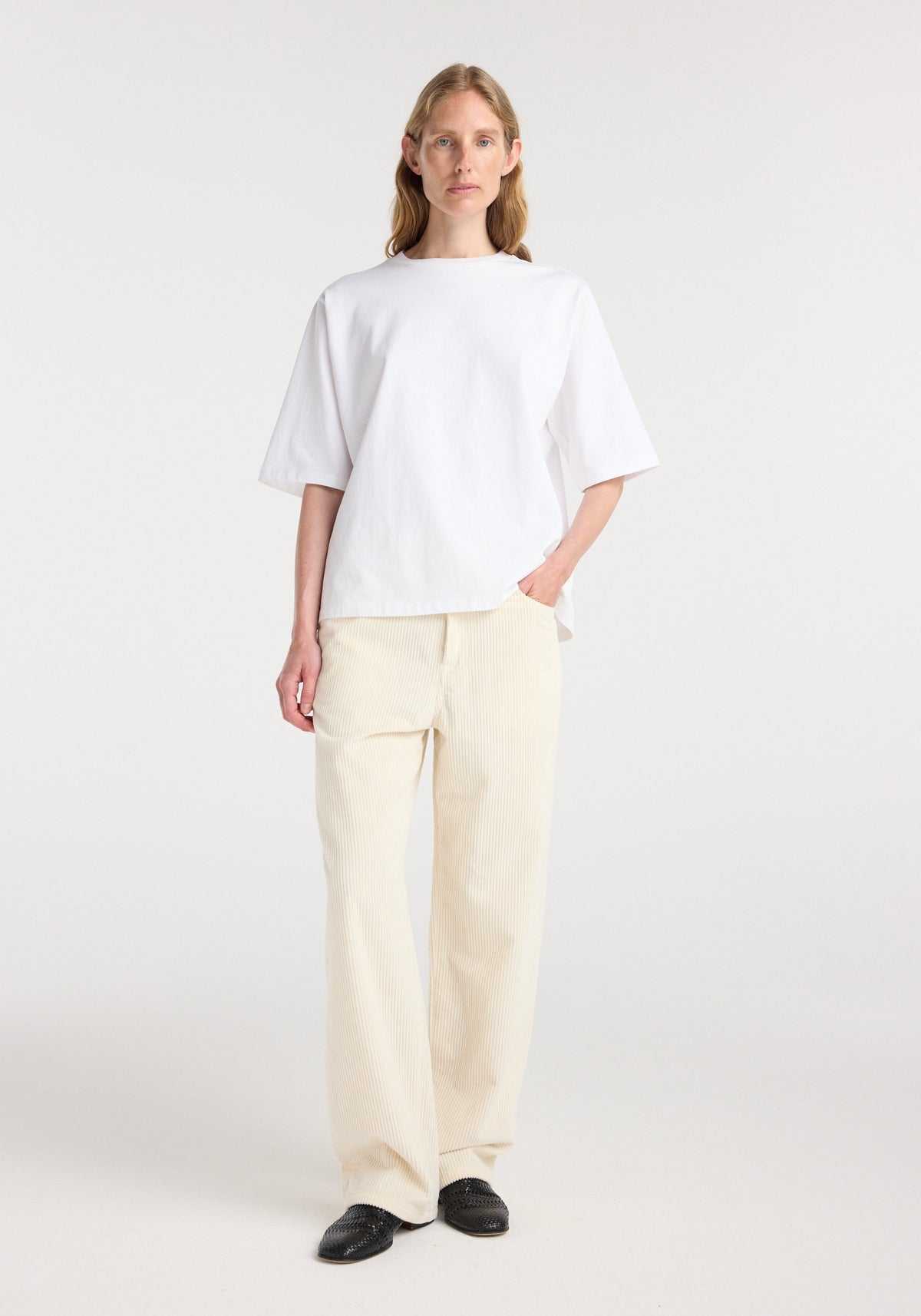 Woman wearing an Oversized cotton t-shirt | optic white with half sleeves paired with light beige wide-leg pants and black slip-on shoes standing against a plain background.