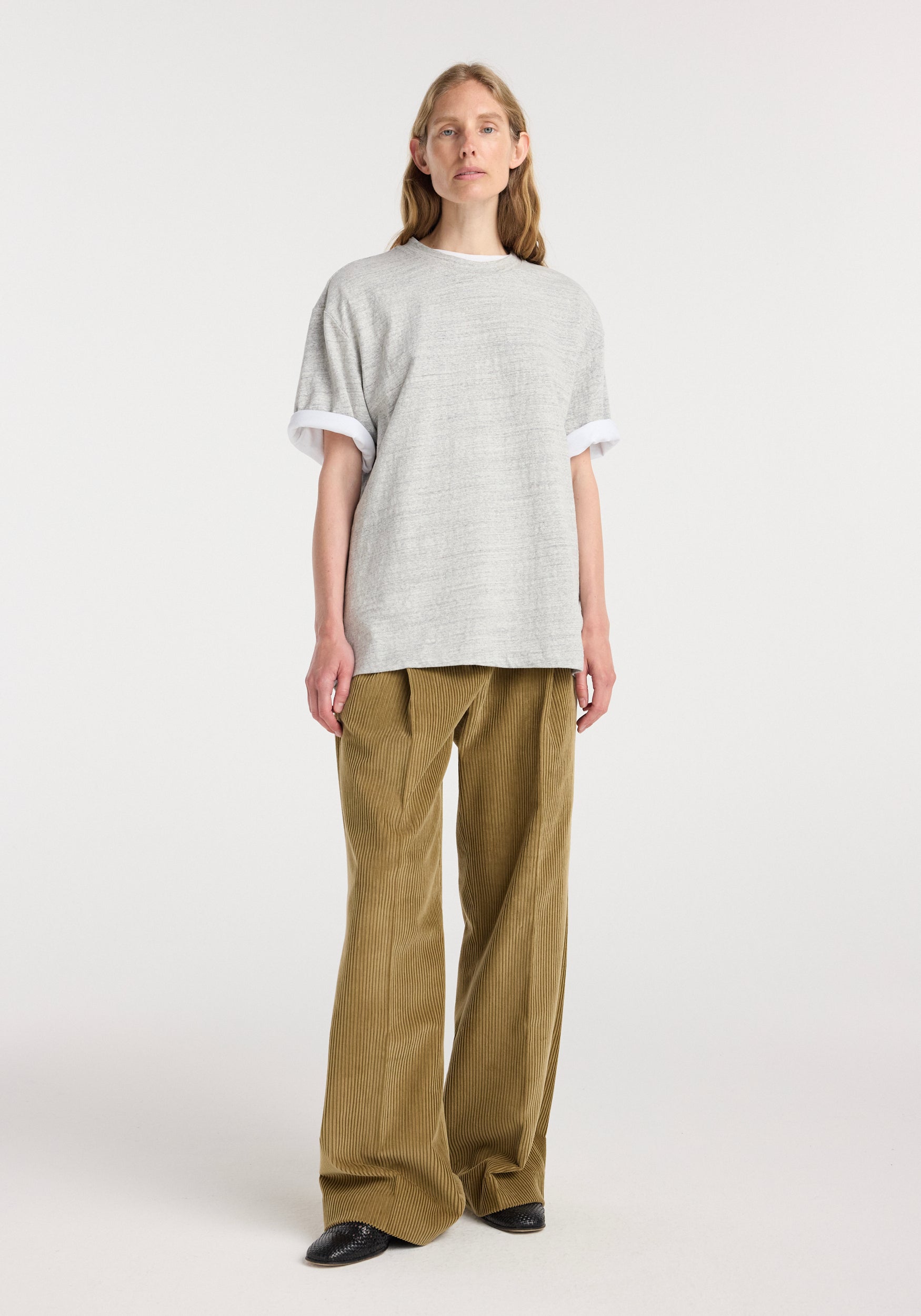 Woman wearing a grey mouliné Japanese mouliné jersey t-shirt with rolled sleeves paired with loose-fit brown corduroy pants and black shoes against a plain white background.