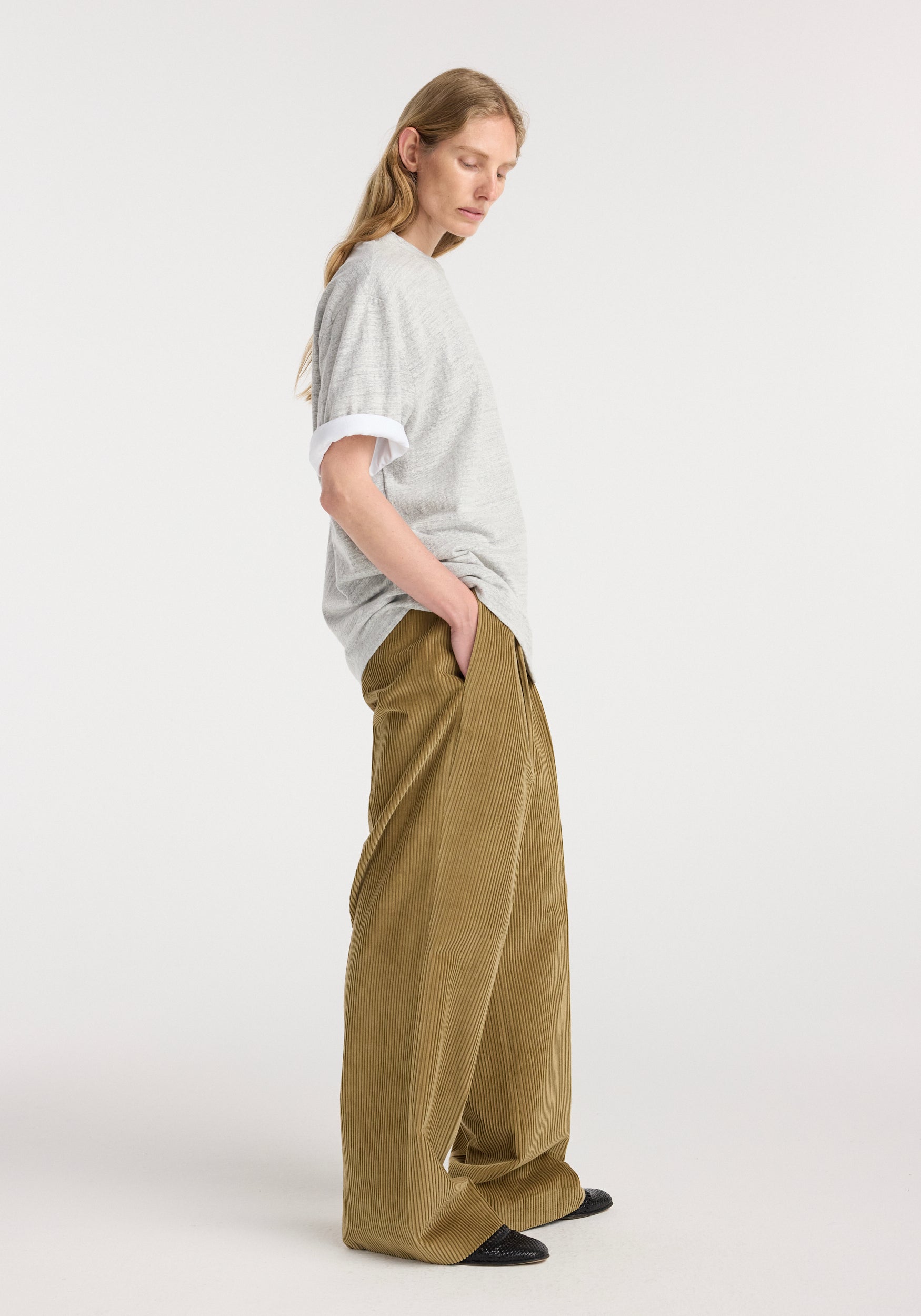 Side view of a woman wearing a grey mouliné Japanese mouliné jersey t-shirt with rolled short sleeves, paired with loose-fitting brown corduroy pants and black shoes, against a plain white background.