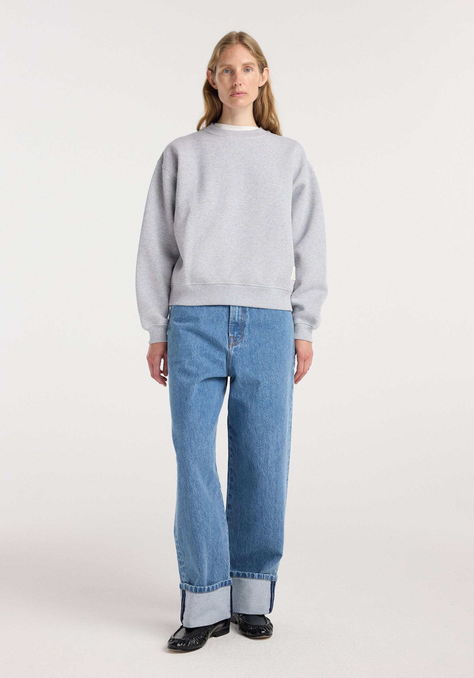 A woman wearing a Sweatshirt | mid grey melange paired with light blue cuffed jeans and black shoes, standing against a plain white background.