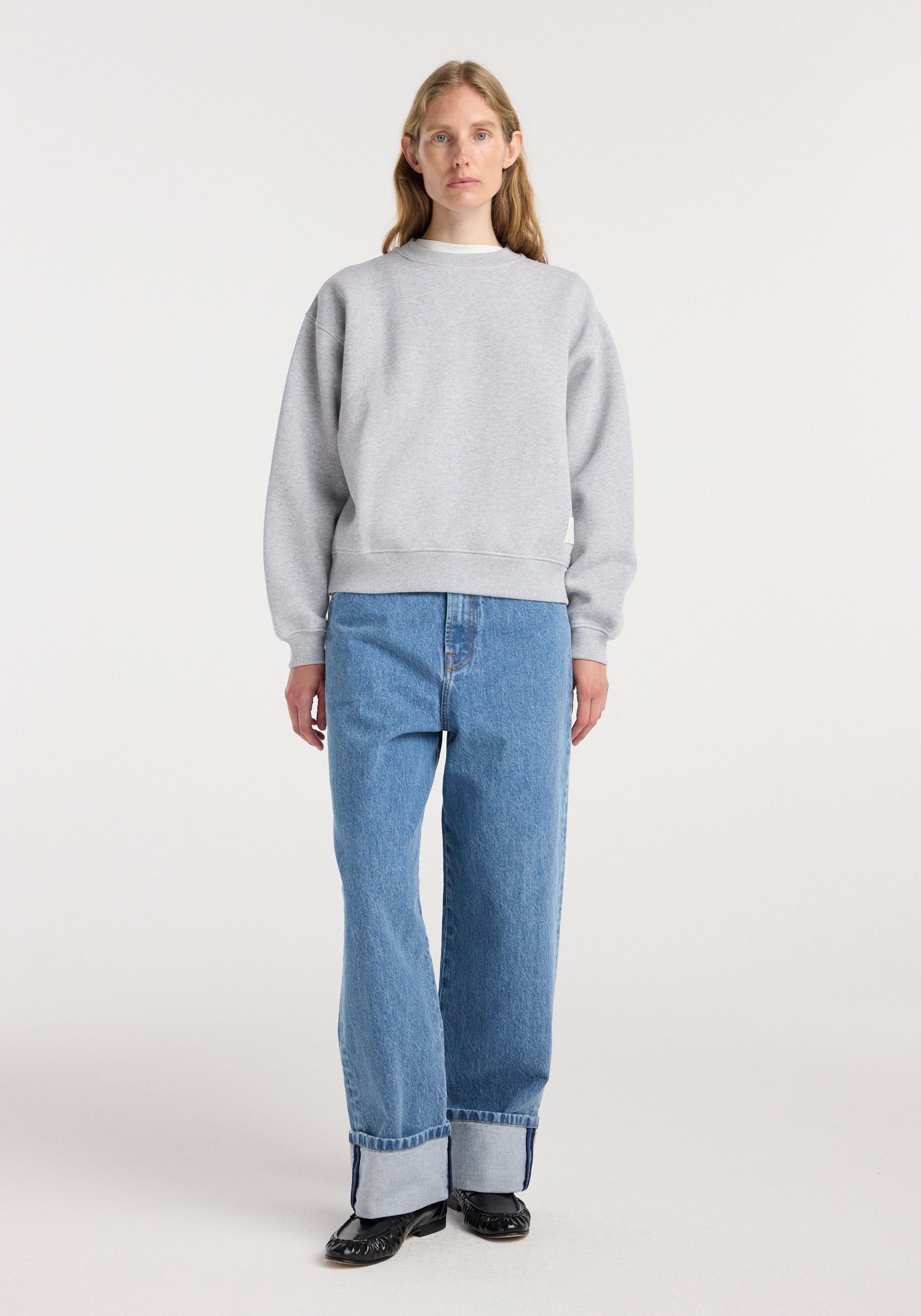 A woman wearing a Sweatshirt | mid grey melange paired with light blue cuffed jeans and black shoes, standing against a plain white background.