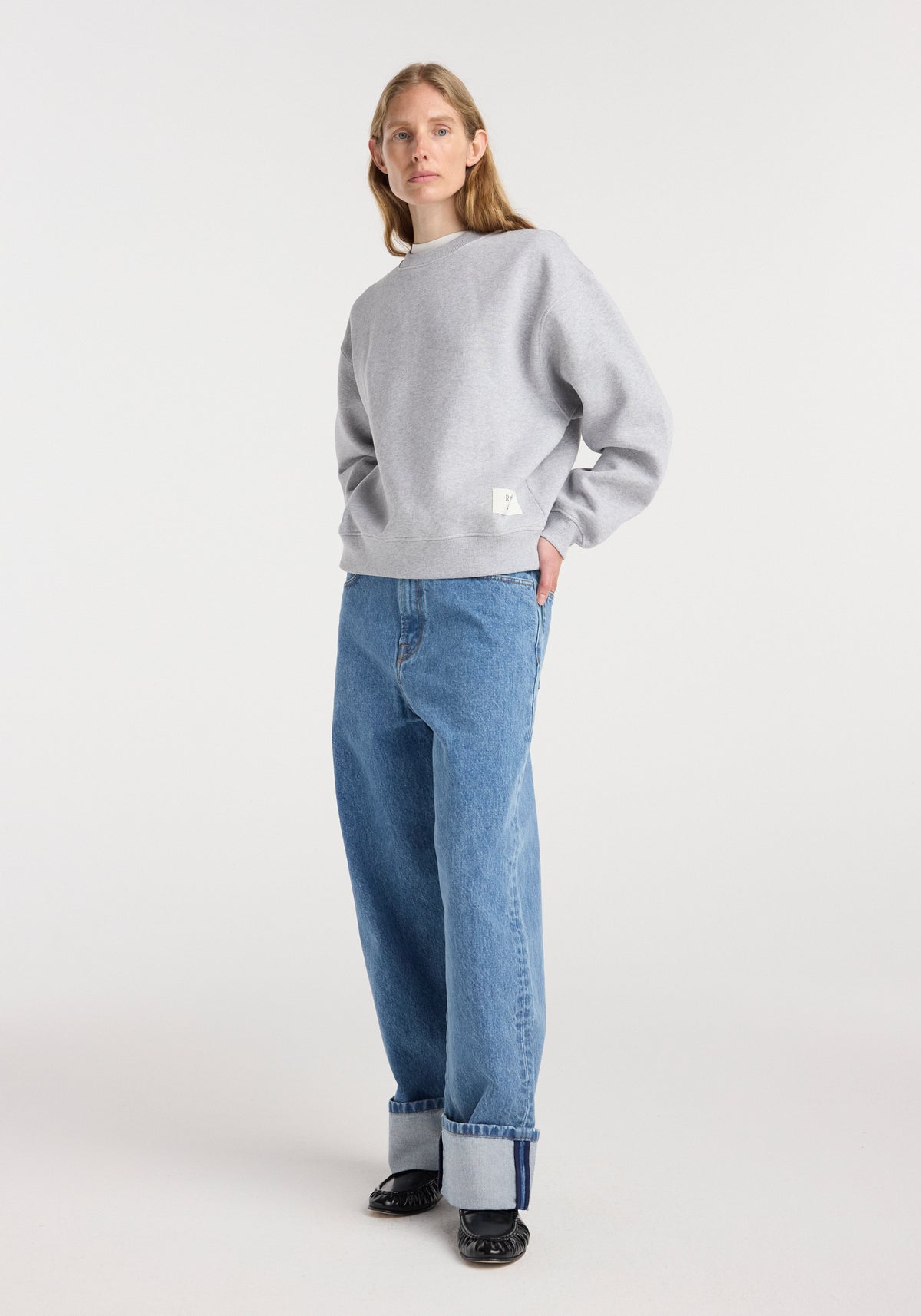 Woman wearing a Sweatshirt | mid grey melange with long sleeves and a relaxed fit, paired with cuffed blue jeans and black shoes, standing against a plain white background.