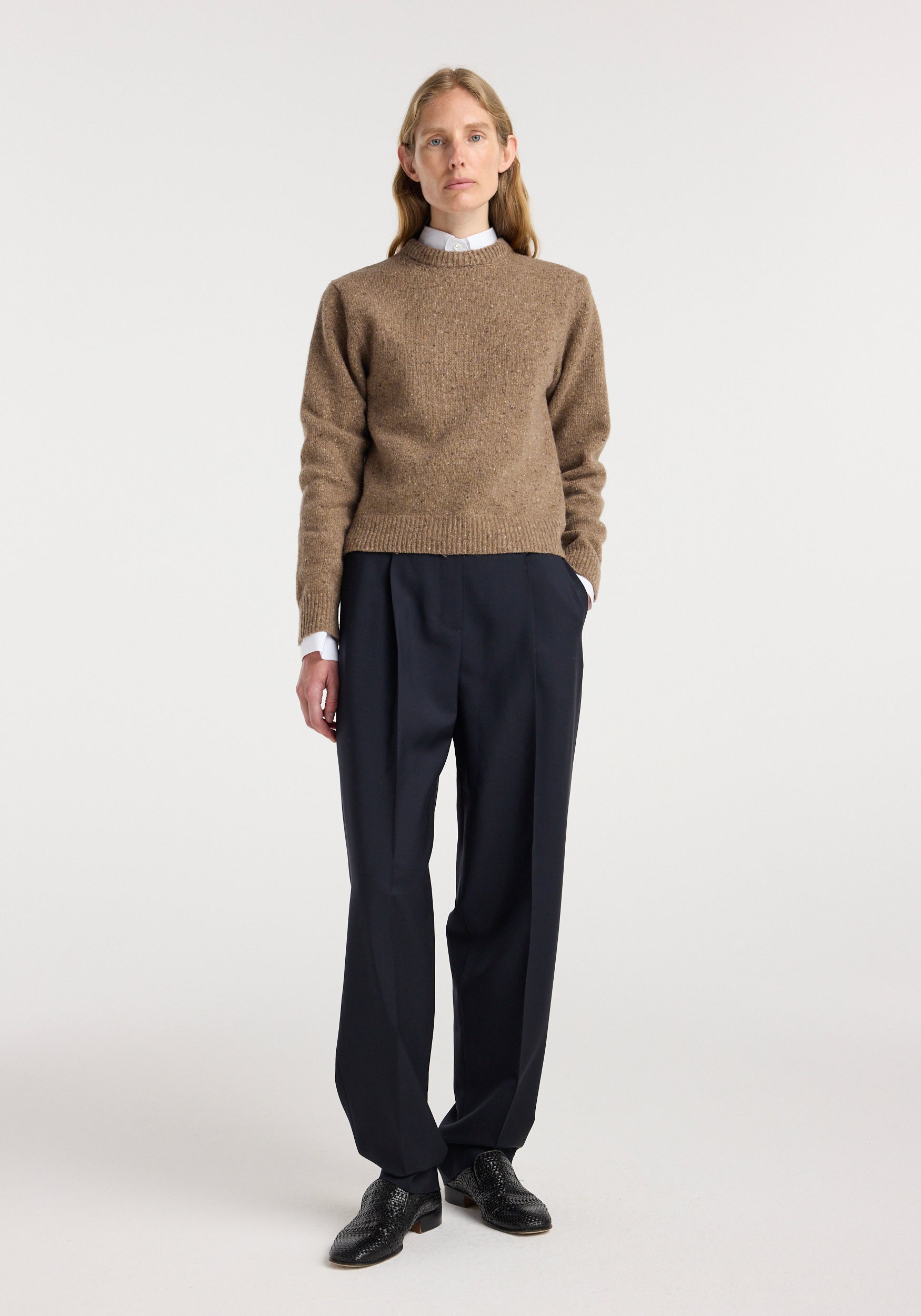 Woman wearing a fitted mouliné crewneck | mouliné taupe melange sweater paired with black pleated trousers and black shoes, standing against a plain background.