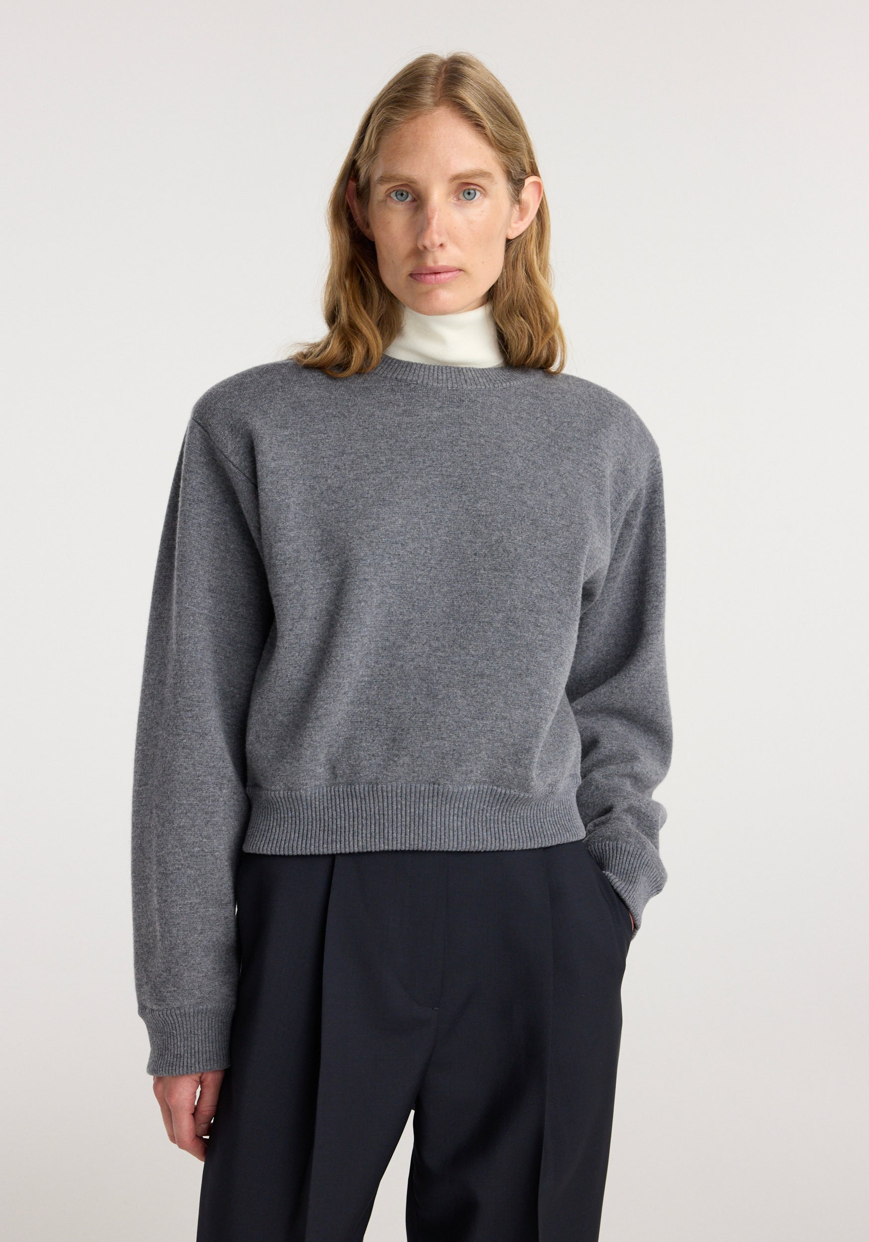 Woman wearing a mid grey melange Statement shoulder crewneck sweater over a white turtleneck, paired with black high-waisted pants, standing against a plain light background.