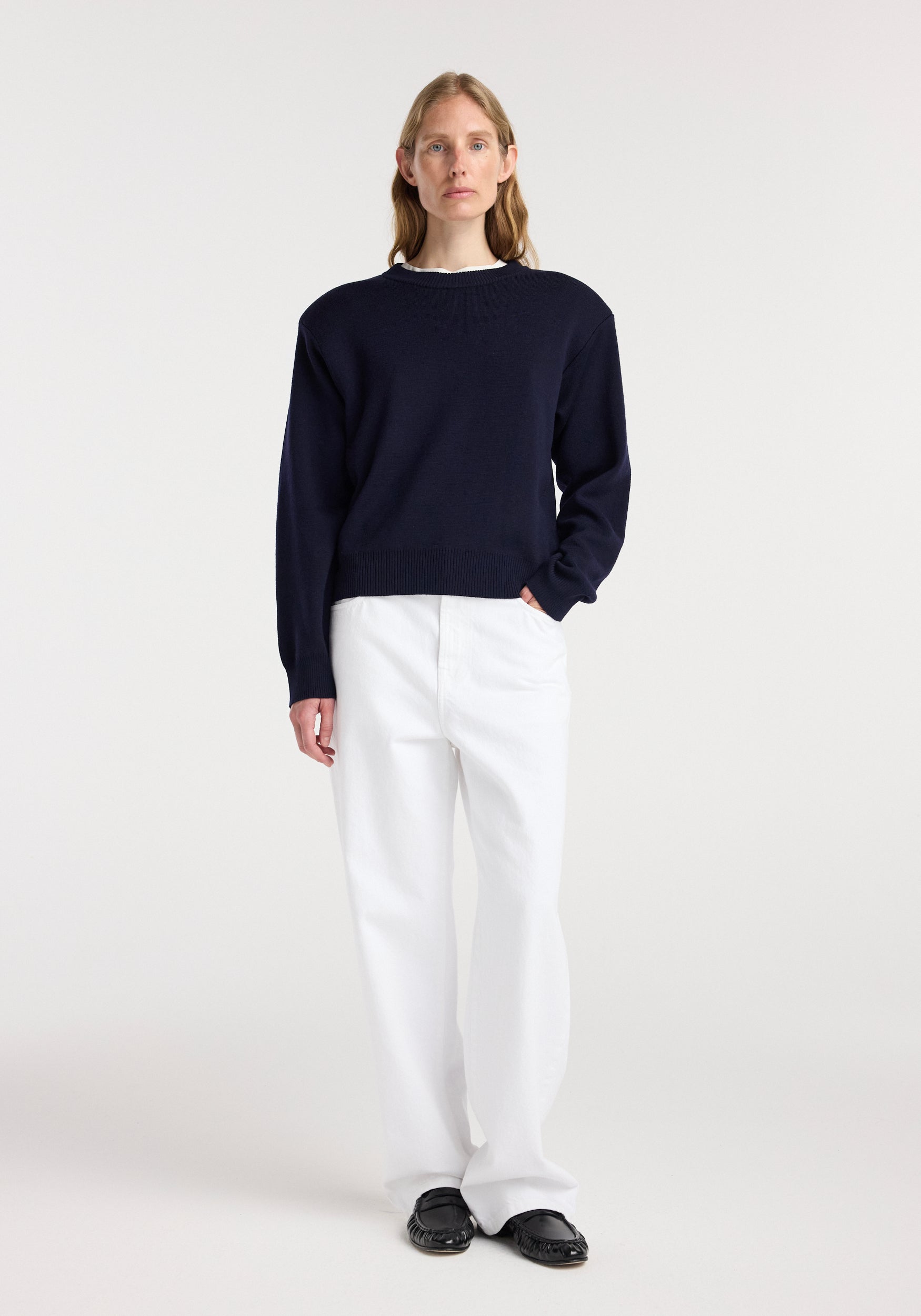 Woman wearing a navy Statement shoulder crewneck paired with white pants and black shoes, standing against a plain light background.