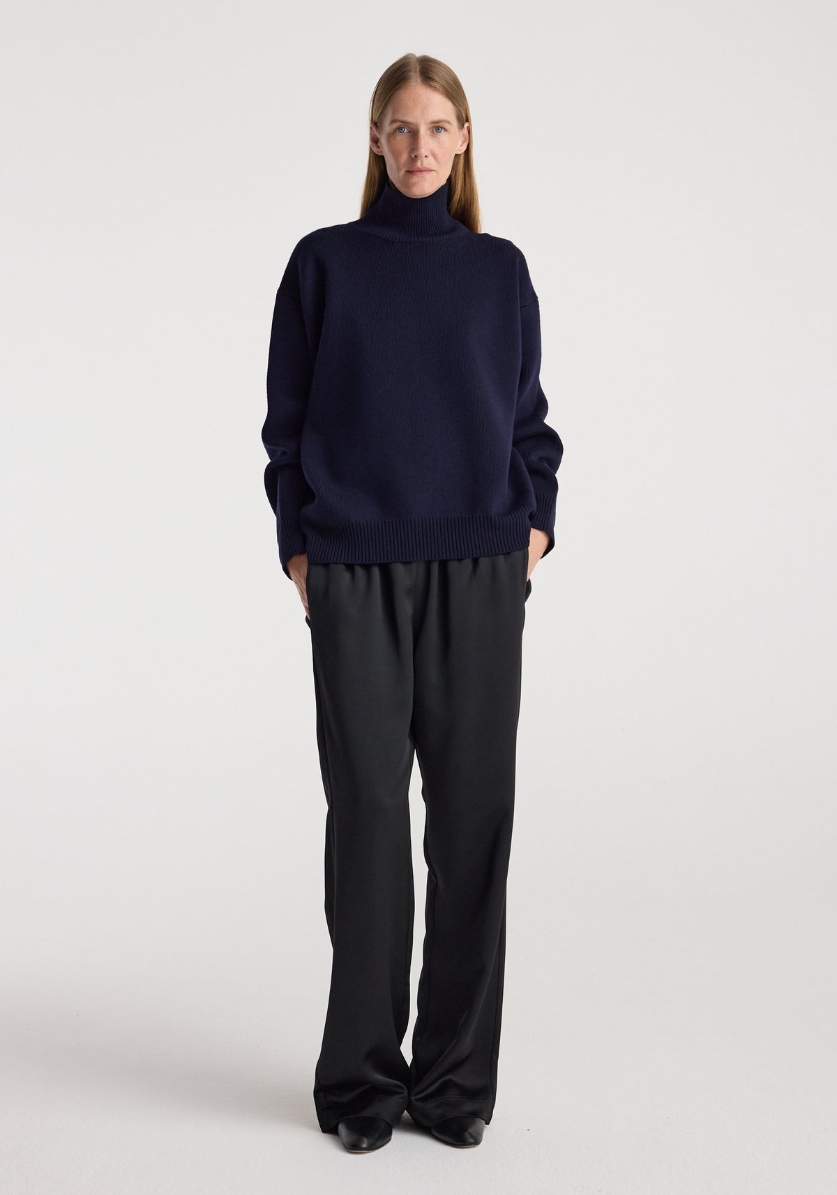Woman wearing a navy Sculptural compact turtleneck | navy sweater with long sleeves, paired with loose black pants and black shoes, standing against a white background.