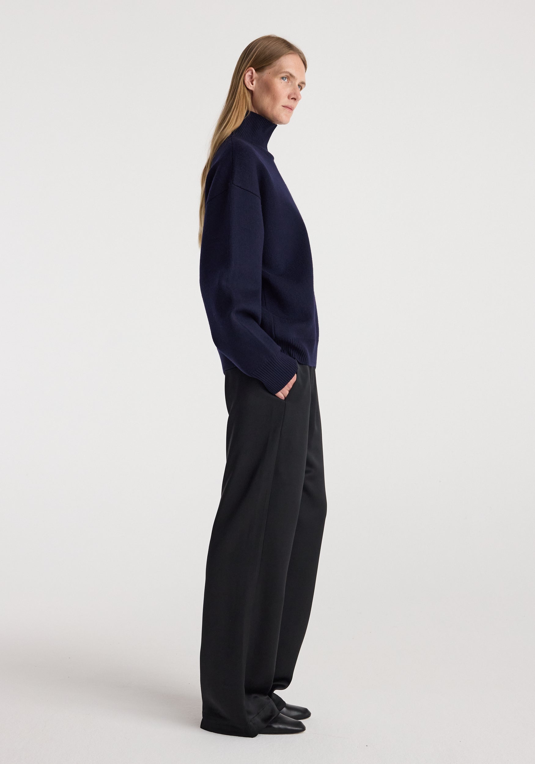 Woman wearing a navy Sculptural compact turtleneck | navy paired with wide-leg black pants and black shoes, standing in profile against a plain white background.